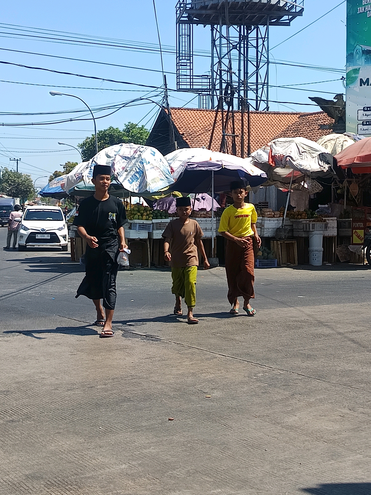 Three young boys, dressed in traditional Islamic attire consisting of peci caps, long shirts, and sarongs, walk across a paved road in front of a market stall. The boy on the left wears a black shirt and black sarong, the middle boy wears a brown shirt and green sarong, and the boy on the right wears a yellow shirt and brown sarong. They are walking in the same direction, with the boy on the right slightly ahead of the other two. In the background, there are market stalls with umbrellas providing shade, displaying fruits and other goods. A white car is parked on the left side of the road. Utility poles and wires are visible in the sky, along with a tall structure with a water tank. The scene appears to be in Blega, Indonesia, under a clear blue sky.