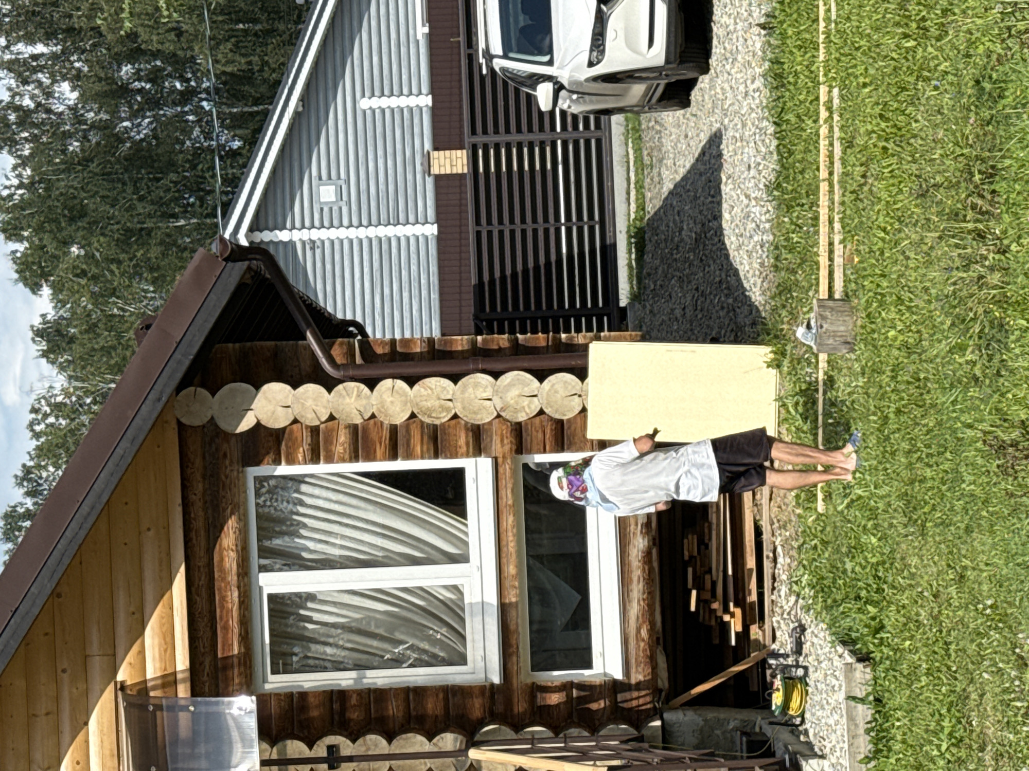 In Kremenkul', Russia, a person stands in a yard, facing away from the camera, near a log cabin. The individual is wearing a white t-shirt, black shorts, and blue flip-flops, with a patterned bandana on their head. They are holding a rectangular object, possibly insulation or a building material, which is yellow in color. To the left of the person are two windows on the log cabin, one with white curtains visible. In the background, another house with horizontal siding is visible, along with a dark metal fence and a white SUV parked on a gravel driveway. The foreground is covered in green grass. The scene appears to be outdoors during the daytime, with dappled sunlight.