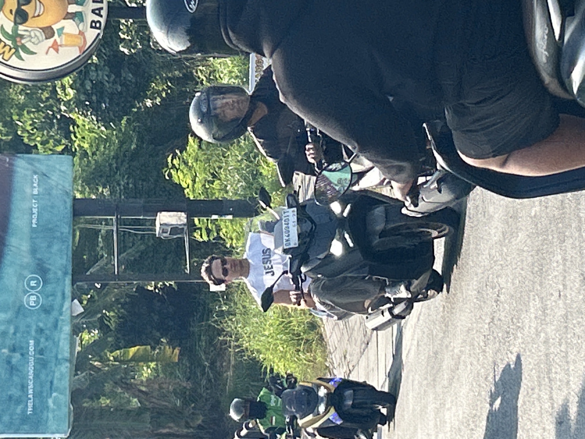 In North Kuta, Indonesia, on a sunny day, a man wearing a white t-shirt with "JESUS" printed on it rides a dark-colored scooter. He has on sunglasses and dark pants. Behind him, another person, wearing a black hoodie and dark shorts, rides a scooter, their face obscured by a helmet. To the left, another individual in a green jacket and a helmet is on a scooter. The background consists of lush green foliage and a bright blue sign overhead with white text. The road surface is asphalt.