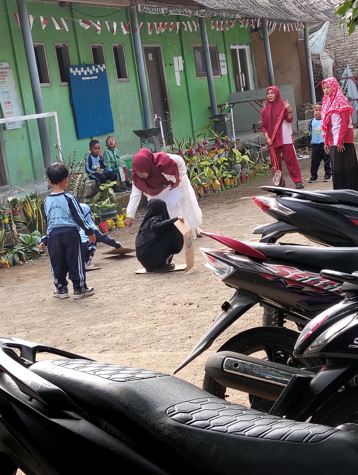 The image depicts an outdoor scene in front of a green building, possibly a school, in Desa Wringin Putih, Indonesia. The building has several windows and is adorned with red and white triangular flags strung across the top. In the foreground, several children are visible. One young boy in a blue and white uniform stands facing away from the camera, looking towards a group of children and adults. Another boy in a similar uniform sits on the ground to his left, looking up. To the right, a woman in a maroon hijab and matching tracksuit holds a long, flat object, possibly a mat or a banner, and appears to be interacting with a child. Another woman in a red and white hijab stands nearby.

The ground is dirt and gravel. In the middle ground, a few children are engaged in an activity involving cardboard. One child, wearing black, is crouched on a piece of cardboard. Another child's legs are visible behind them. In the background, several motorcycles are parked. The environment suggests a schoolyard or community event. The overall atmosphere appears to be casual and participatory.