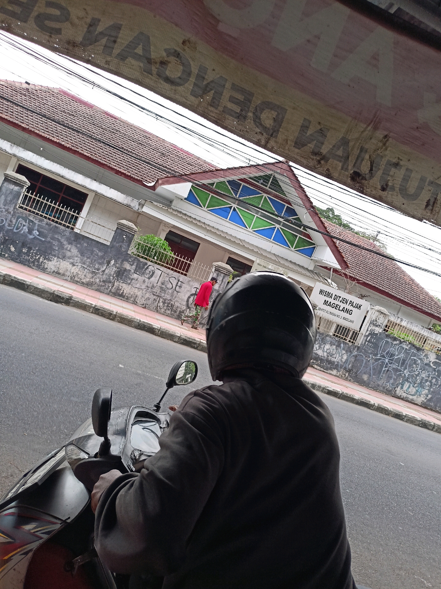 The image shows a view from the perspective of someone riding a scooter in the city of Magelang, Indonesia. The person is wearing a dark helmet and a dark long-sleeved shirt. In front of them, a building with a tiled roof is visible, with a sign that reads "WISMA DJTJEN PAJAK MAGELANG" and further text below it. A sidewalk runs in front of the building, with a person in a red shirt walking on it. The road is paved and appears to be a city street. There are overhead wires visible in the frame. A blurred sign with text is visible at the top of the image.