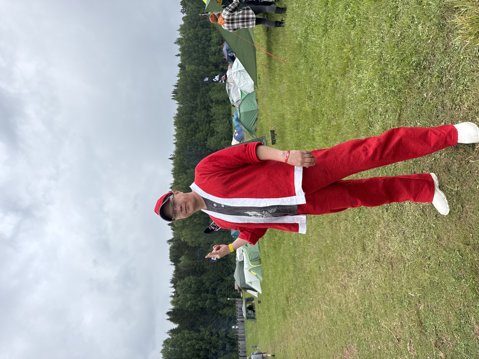 The image captures an outdoor scene in a grassy field, identified as Bol'shaya Kivara, Russia, under an overcast sky. A man of apparent East Asian ethnicity, estimated to be middle-aged, stands prominently in the foreground, facing slightly left of the camera with a direct gaze. He is dressed in a red two-piece suit with white trim, resembling a lightweight Santa costume, worn over a grey t-shirt with a faded graphic design. He wears clear-rimmed eyeglasses, a red baseball cap, a yellow wristband on his right wrist, a red wristband on his left wrist, and white slip-on shoes. His right arm is raised, with his index and pinky fingers extended and the middle and ring fingers curled, forming a "rock on" or similar gesture.
The background reveals a large, informal encampment consisting of numerous tents of various colors (predominantly green, grey, and white, with some blue and dark tents) spread across the field. Several tents are partially covered with tarps, and red tent ropes are visible. To the far left, a person in dark clothing walks away from the camera, with a smaller figure following behind them. Further left, behind the tents, a wooden fence or structure is visible, with a faint plume of smoke or steam rising beyond it. To the right, another person is seen from behind, wearing a plaid shirt, an orange beanie, and dark pants, standing near a large green tarp setup. A dense line of mixed coniferous and deciduous trees forms the distant horizon line. The ground is a mix of green grass and patches of worn earth. The sky is uniformly grey with thick clouds, indicating an overcast day.