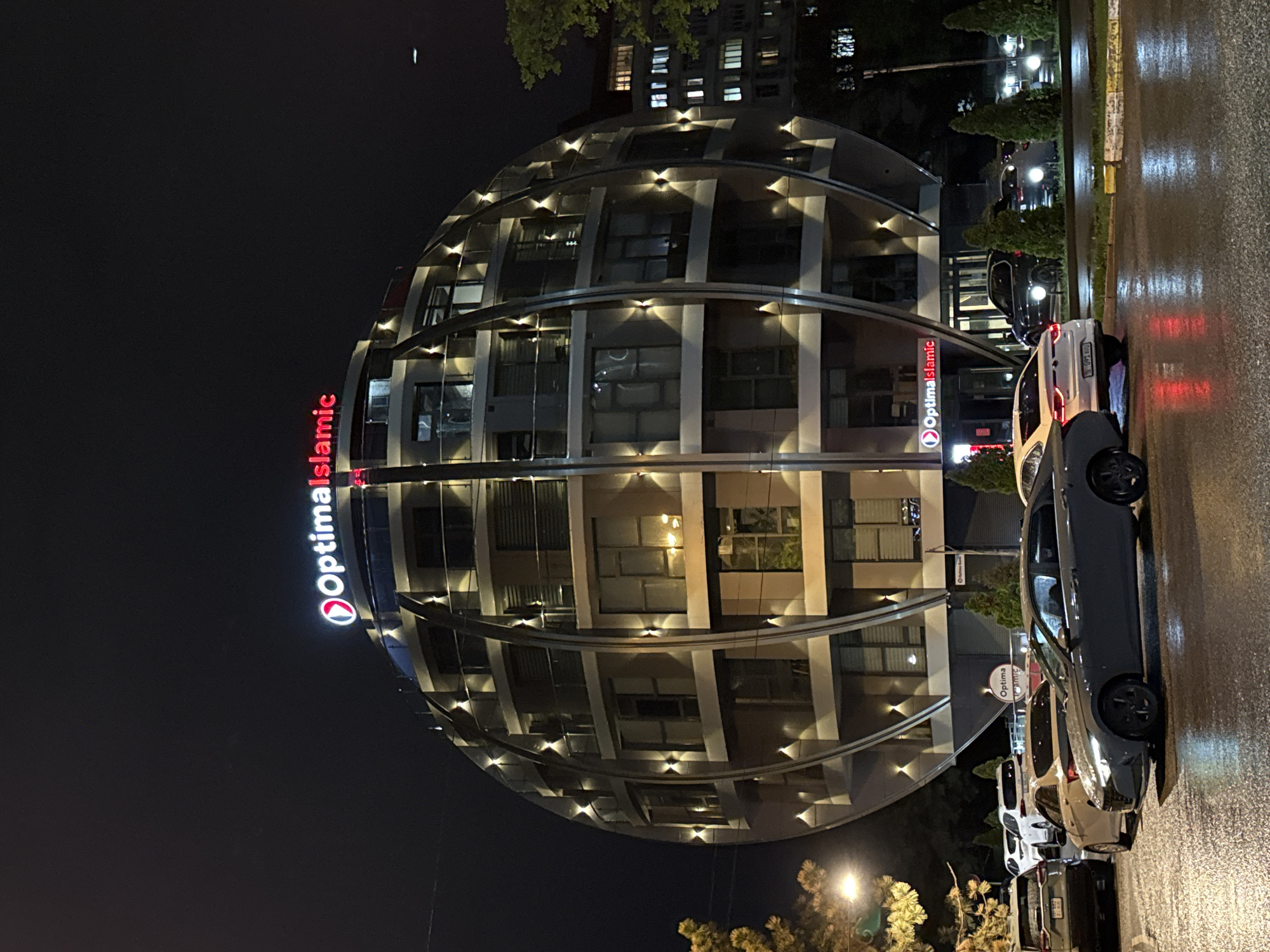 The image captures an urban night scene in Bishkek City, Kyrgyzstan, dominated by a distinctive, brightly lit building. The building features a unique spherical or domed architectural design, clad in light-colored panels and extensive glass windows. Its curved exterior is illuminated by numerous sconce-style lights arranged in horizontal rows, emphasizing its modern aesthetic.
Two prominent signs for "Optima Islamic" are visible on the building's facade. One large sign, with red lettering and a white background, is oriented vertically on the left side of the building, while a smaller, similarly branded sign is located on the right side near the street level.
The setting is an urban street or parking area at night. The ground is visibly wet, reflecting the artificial lights from the building and streetlights, suggesting recent rainfall. Several cars are parked along the curb, including a dark-colored sedan prominently featured in the foreground on the right, a white hatchback, and other vehicles further down the street. Overhead, the sky is dark, with a faint light source visible in the upper left corner. Lush green trees are scattered around, their foliage partially illuminated by nearby light sources. In the background, other multi-story buildings with illuminated windows can be seen, contributing to the urban nighttime atmosphere. No human activity is discernable, creating a still and illuminated cityscape.
