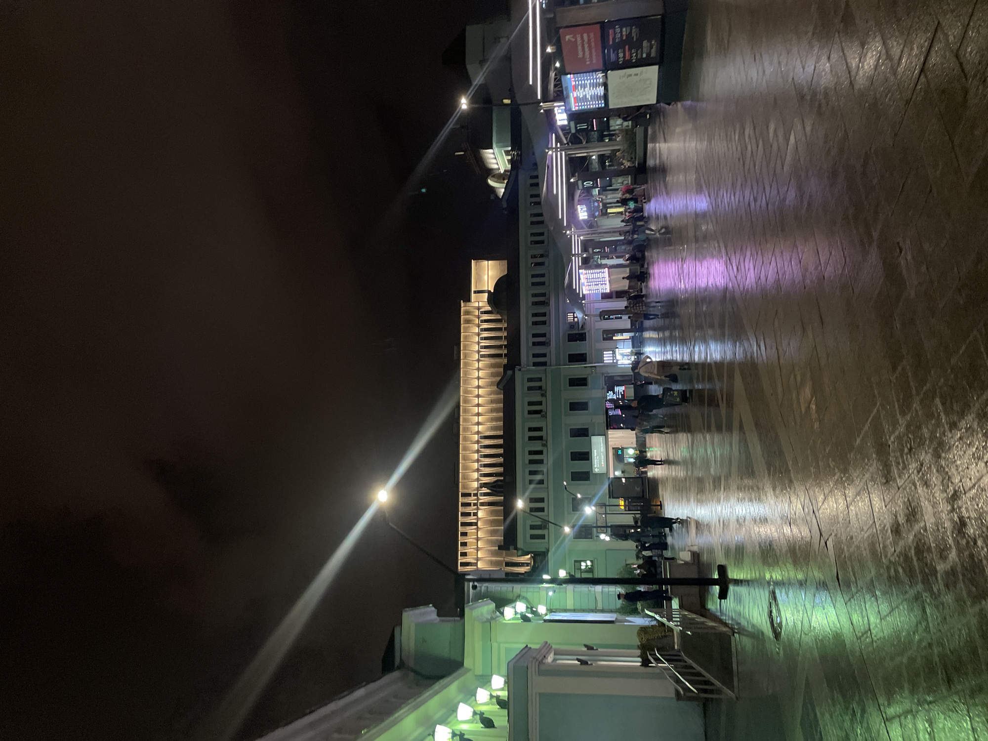 The image depicts a nighttime view of a station square, likely a train or bus station, in Moscow, Russia. The ground is wet and reflects the colorful lights from the surrounding buildings and signage, creating a glistening effect. Several people are visible walking through the square, some appearing to be travelers with luggage. Modern digital displays showing information, possibly train or flight schedules, are present, indicating a busy transportation hub. The architecture of the buildings suggests a blend of historical and contemporary design, with illuminated facades and decorative lighting enhancing the nighttime ambiance. The sky above is dark and cloudy, with some light beams from streetlights cutting through the night.