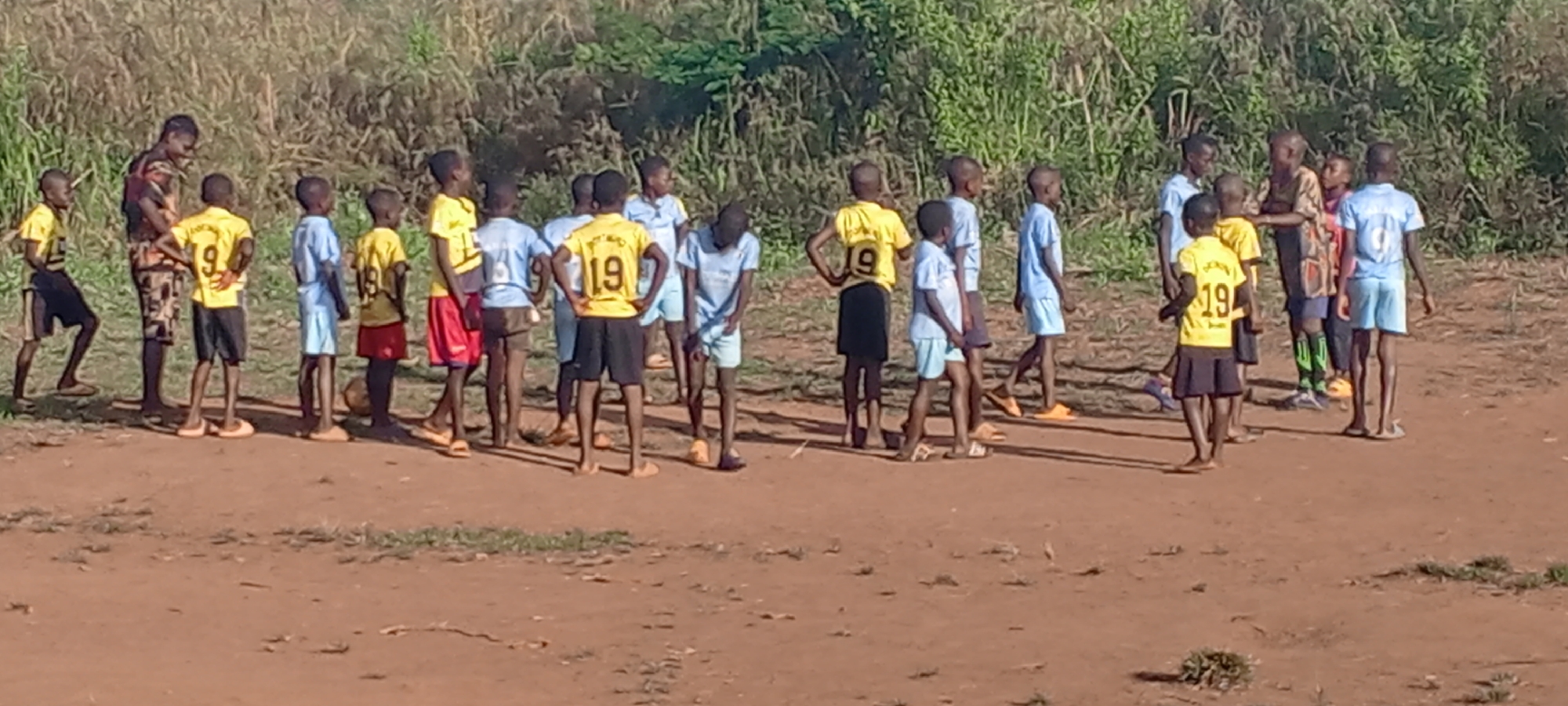 This image depicts a group of approximately twenty children and a few adults gathered on a reddish-brown dirt field. The background is characterized by dense green foliage and trees, indicating an outdoor, natural setting in Ngalula, Democratic Republic of the Congo.
The children, mostly young boys, are wearing sports jerseys. Some are in yellow shirts with visible numbers such as "9" and "19," while others wear light blue shirts, also with numbers like "9." Many are barefoot or wearing simple sandals/flip-flops. One adult figure, dressed in a patterned shirt and shorts, is prominent on the left, interacting with a cluster of children. Another adult is visible further to the right.
The grouping of children and adults, coupled with the sports attire and the faint presence of a soccer ball near one child's feet, suggests a football (soccer) practice, training session, or a team gathering. The lighting indicates a sunny day, likely late morning or early afternoon, with relatively short shadows on the ground. The overall mood appears calm and focused.