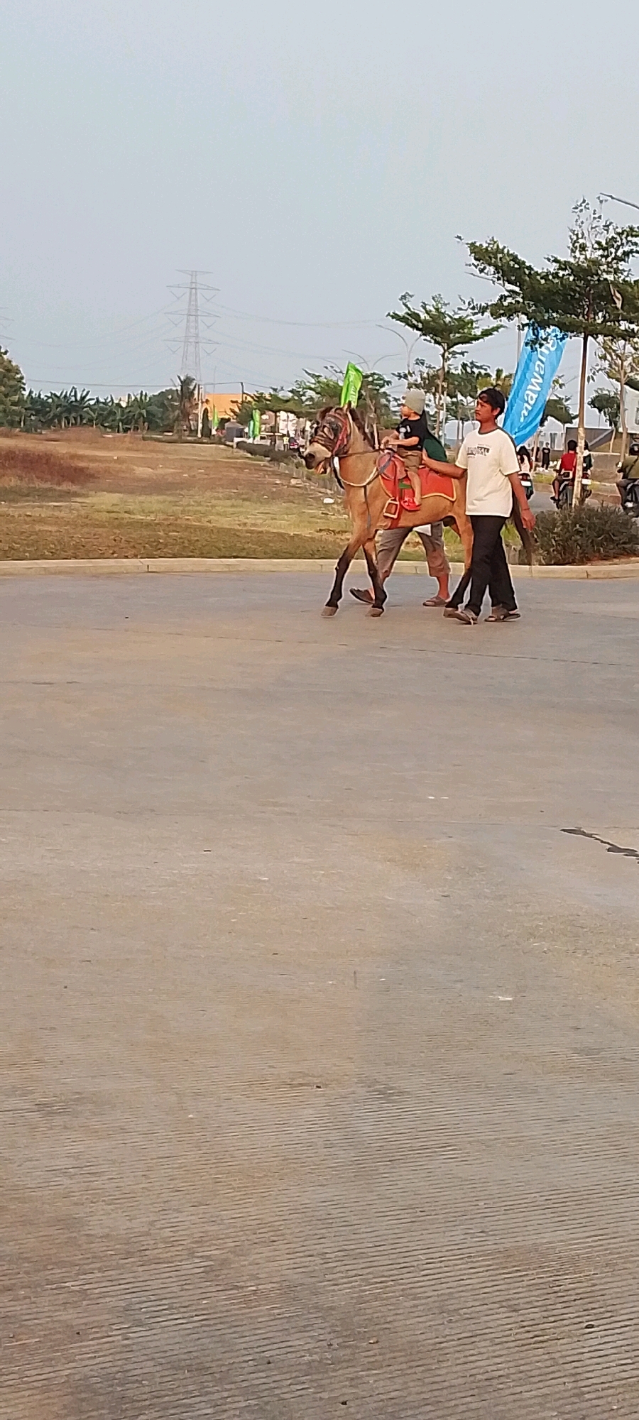 In Satriamekar, Indonesia, a young boy in a light-colored hat and a dark shirt rides a light brown horse. He is seated on a red saddle pad. A young man, wearing a white t-shirt with text and dark pants, walks beside the horse, holding its reins. Another person, partially visible, stands behind them, also appearing to be involved with the horse. The horse is walking on a paved surface. In the background, there is a grassy field with some trees and a power tower. There are also banners and some people and vehicles further in the distance. The sky is overcast.
