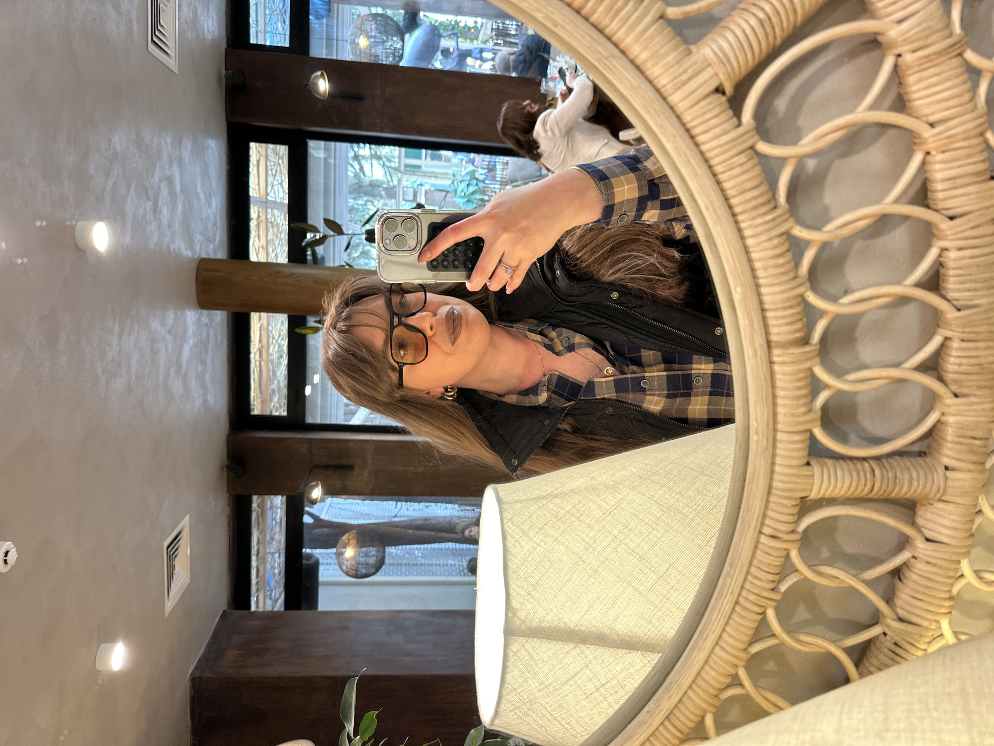 A woman with blonde hair and glasses is taking a selfie in a round rattan mirror in a public place, likely a restaurant or cafe. She is wearing a plaid shirt and a black vest. In the background, there are other people and windows. The location is presumed to be Tbilisi, Georgia.