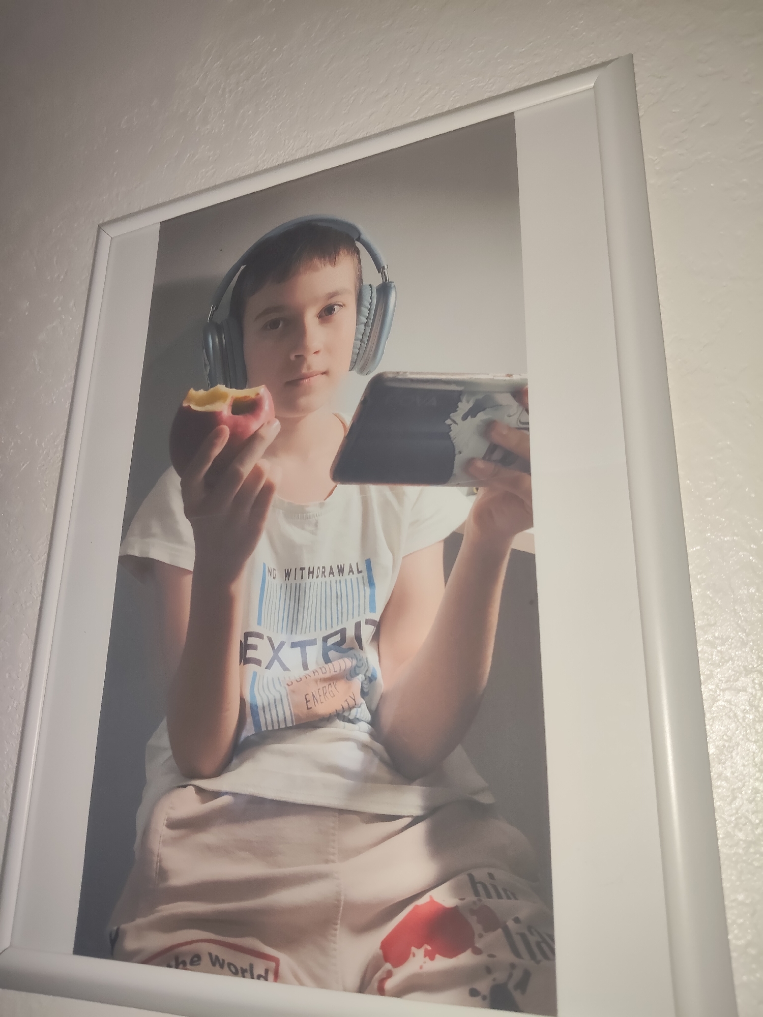 The image is a framed photograph of a young boy wearing blue headphones. He is holding a bitten apple in his left hand and a tablet in his right hand. The boy is wearing a white t-shirt with blue and white text that reads "NO WITHDRAWAL" and "EXTRI." Below this, the text "ENERGY" is visible. He is also wearing light-colored shorts with red and black graphics, including the words "the World" in red lettering. The photograph appears to be taken indoors, with a plain, light-colored wall as the background. The frame is white. The image does not contain any specific landmarks or environmental cues that would allow for precise identification of its location within Reftinsky, Russia.