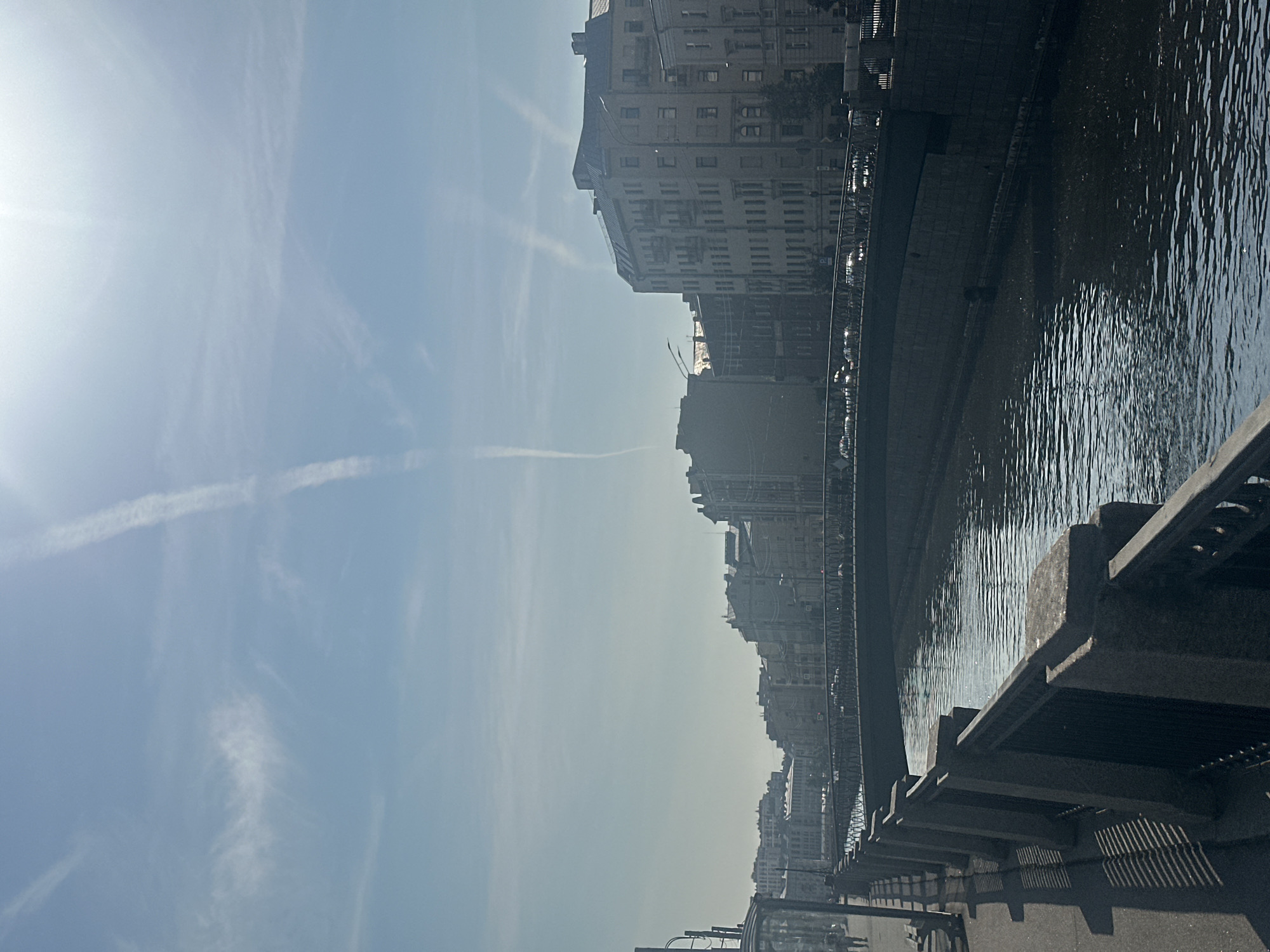 An eye-level, full outdoor, medium shot captures a tranquil canal flowing through a European city under a bright, hazy sky. Contrails streak across the pale blue expanse, with the sun glinting at the top right, casting a soft glow on the scene. The canal is bordered by stone embankments, with dark, ornate metal railings lining the walkway on the left. Across the water, a row of historic, multi-story buildings with light-colored facades and numerous windows stretches into the distance. A pedestrian bridge spans the canal, with vehicles visible crossing it. The architecture suggests a city with a rich history, possibly St. Petersburg, Russia, given the canal and building styles. The atmosphere is serene, with the gentle ripples on the water reflecting the overcast sky.