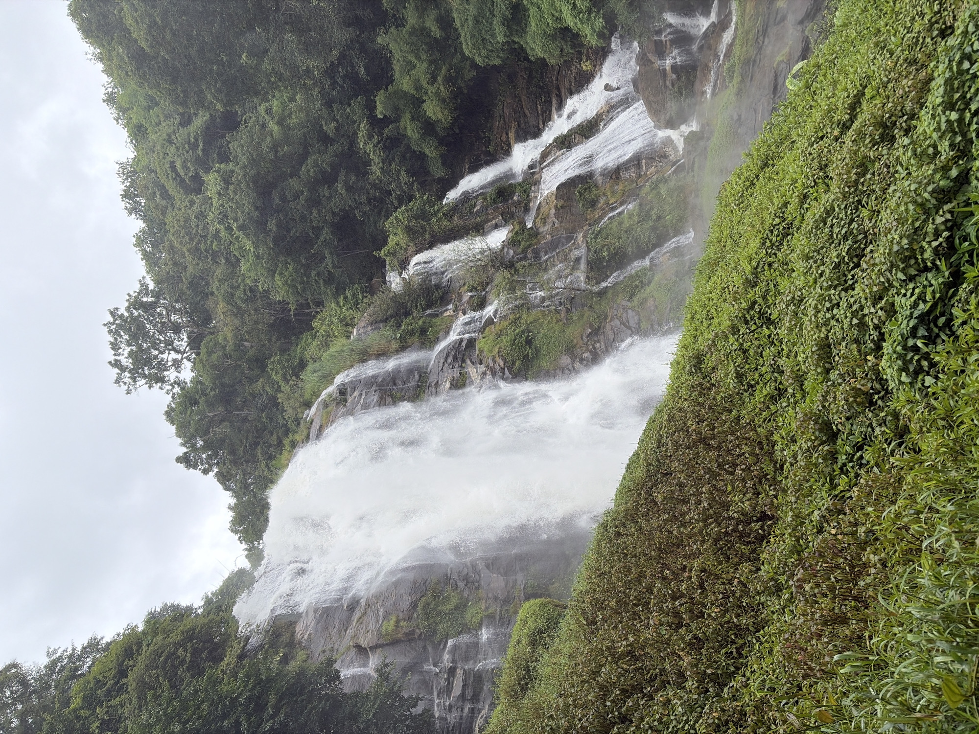 A wide, multi-tiered waterfall cascades down a rocky, moss-covered cliff face, surrounded by lush green vegetation. The water is a bright white, with a fine mist rising from its impact. The sky above is overcast, with a pale grey hue. The scene appears to be a natural landscape, likely in a tropical or subtropical region, given the dense foliage. The location is identified as Ban Sop Hat, Thailand.