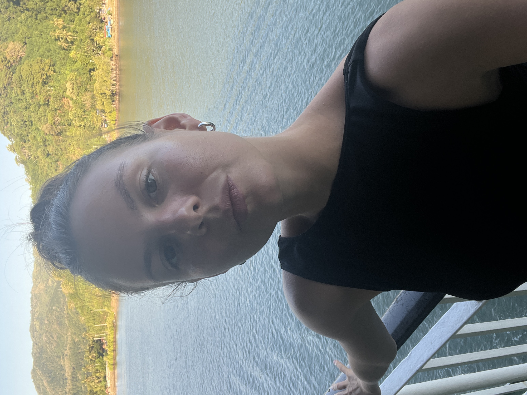 A close-up, eye-level selfie shows a woman with her hair pulled back in a bun, looking directly at the camera. She is wearing a black sleeveless top and a silver hoop earring in her right ear. Her left hand is resting on a white railing. In the background, there is a body of water and a lush green hillside with palm trees and some buildings. The scene appears to be set in Batas Desa Skotong Timur, Indonesia, likely on a ferry or boat as indicated by the railing and the open water. The sun is shining, casting a warm light on the scene.