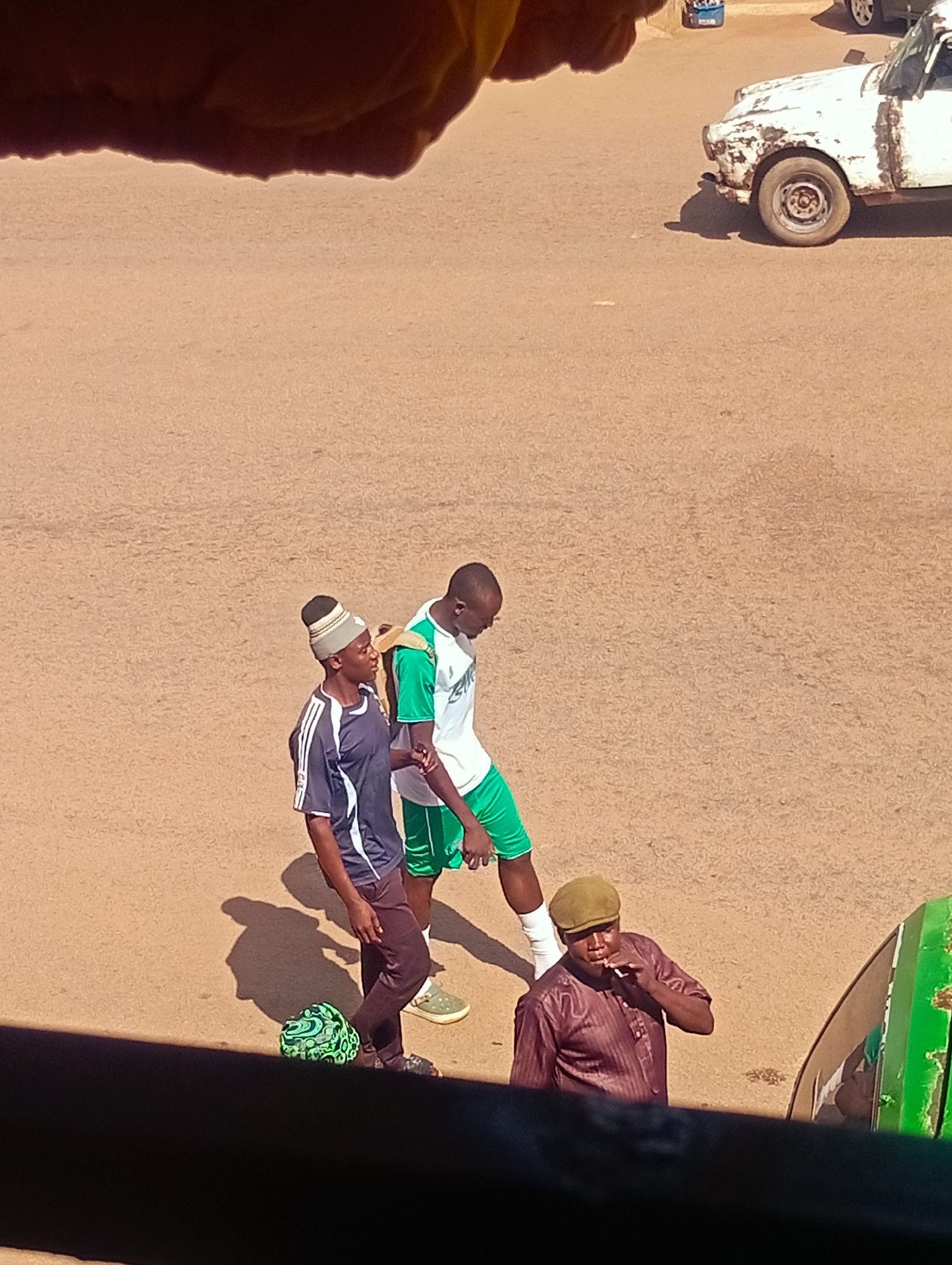In Bukuru, Nigeria, a street scene unfolds with three individuals. Two young men, one wearing a striped beanie and a dark blue t-shirt with white stripes on the sleeves, and the other in a green and white athletic shirt, walk side-by-side. The former has a soccer ball with a green and black pattern at his feet. Behind them, an older man in a maroon striped shirt and a brown cap smokes a cigarette. A dilapidated white vehicle is parked on the right side of the frame, its body showing significant rust and damage. The ground is a sandy, reddish-brown surface, indicative of a dusty street. A portion of a green vehicle is visible at the bottom right edge of the image. The lighting suggests it is daytime, with shadows cast on the ground.