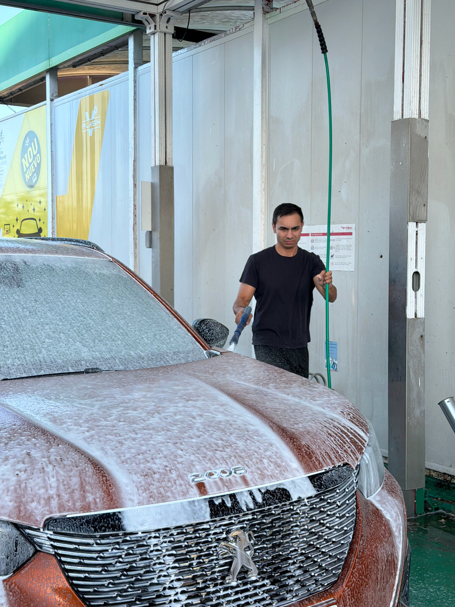 In Cambrils, Spain, a man is washing a car at a self-service car wash. The car, a red Peugeot 2008, is covered in white foam. The man, wearing a black t-shirt and black pants, is holding a green hose with one hand and a blue wash brush with the other, appearing to be in the process of rinsing the vehicle. The car wash bay has white paneling walls and a grey metal structure on the right. A yellow and white sign with the word "FLASH" is visible in the background, and another sign with text in Spanish is mounted on the wall behind the man. The ground is wet and green.