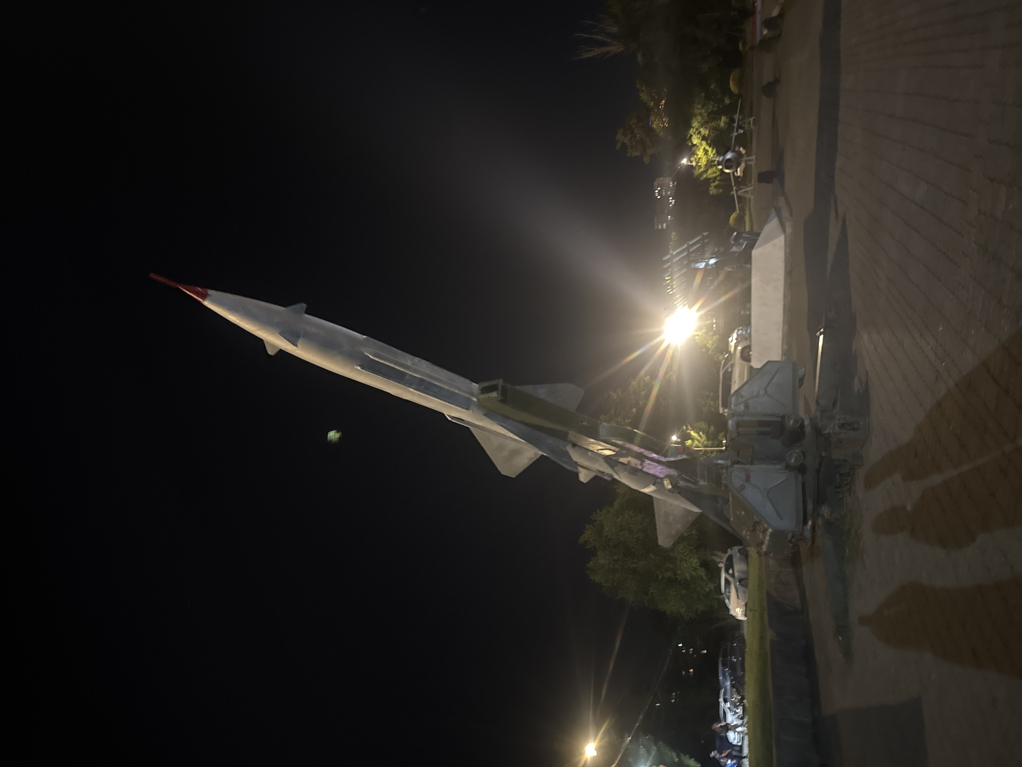 The image captures an outdoor night scene within Victory Park in Yerevan, Armenia. A prominent, static display of a large, grey surface-to-air missile, identifiable as an S-75 Dvina (SA-2 Guideline) type, stands angled upwards towards the upper right, featuring a distinct red nose cone and mounted on a grey launch platform. To the right of the missile, further in the mid-ground, a grey jet fighter aircraft is visible on display.
The area is artificially lit, with bright streetlights producing significant lens flares on both the left and right sides of the frame. In the left mid-ground, beyond a treeline, several parked white vehicles and indistinct human figures are present. The foreground consists of a paved surface, likely interlocking pavers, exhibiting long, dark shadows of at least two individuals stretching from the lower left across the ground. The sky is dark, indicating nighttime conditions, with faint city lights visible in the distant background. A small, blurry green object is minimally visible in the upper right portion of the sky.