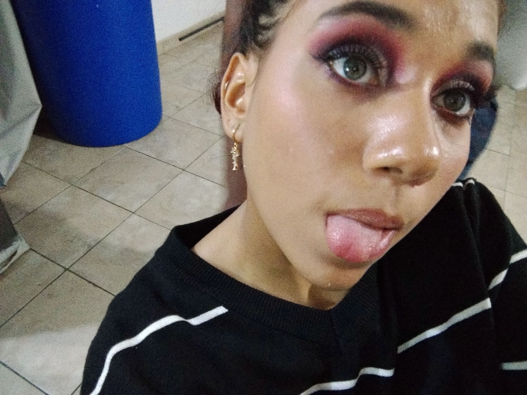 A medium shot shows a young woman sticking her tongue out, her eyes wide and looking toward the camera. She is wearing a black sweater with white stripes and has on dramatic makeup, including pink eyeshadow and long lashes. A gold earring hangs from her left ear. The background appears to be an indoor space with a tiled floor and a large blue cylindrical object. The scene is located in Parroquia Charallave, Venezuela.