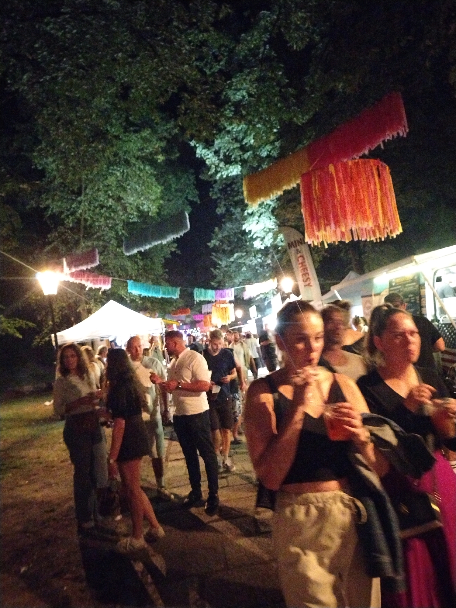 The image depicts an outdoor evening event, likely a festival or market, in Písek, Czechia. Several people are visible, walking and congregating. In the foreground, two women are holding drinks. Behind them, a cluster of individuals appears to be interacting. Tents and food stalls are present, suggesting commercial activity. Overhead, decorative elements, possibly streamers or banners, are suspended. Trees and ambient lighting contribute to the nighttime atmosphere.