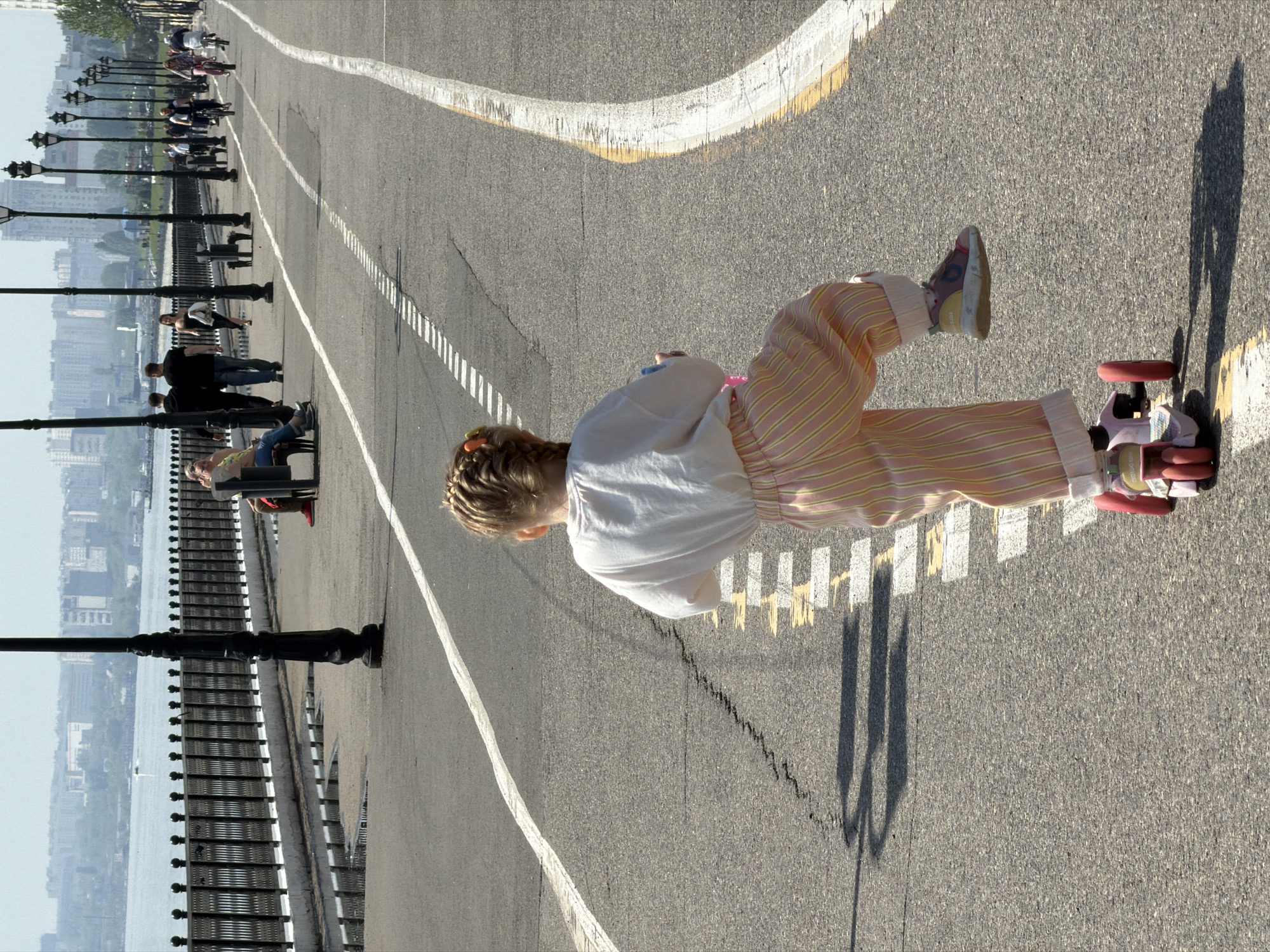 A young girl with blonde braided hair is riding a pink scooter on an asphalt path. She wears a white shirt and striped pink and yellow pants, and multi-colored shoes. The path has white and yellow painted lines. In the background, people are sitting on benches next to black lampposts, and a city skyline is visible across a body of water. This scene is likely in Saratov, Russia, based on its features.
