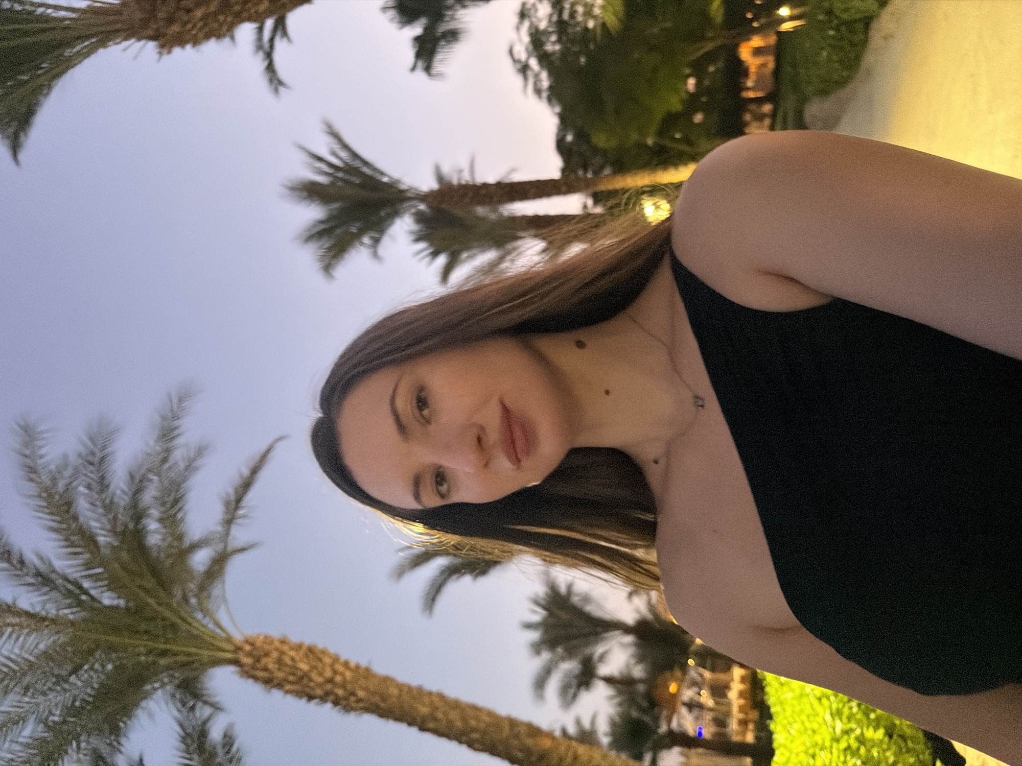 A woman with long, dark hair is taking a selfie outdoors. She is wearing a black, one-shoulder top and a delicate necklace. Her skin is fair, and she has subtle makeup on. Behind her, several palm trees are visible against a twilight sky, suggesting a tropical or desert environment, consistent with locations like Sharm El Sheikh, Egypt. The lighting indicates it is evening, with some artificial lights glowing in the background. The woman is positioned in the foreground, with the natural and man-made elements of the environment forming the backdrop.
