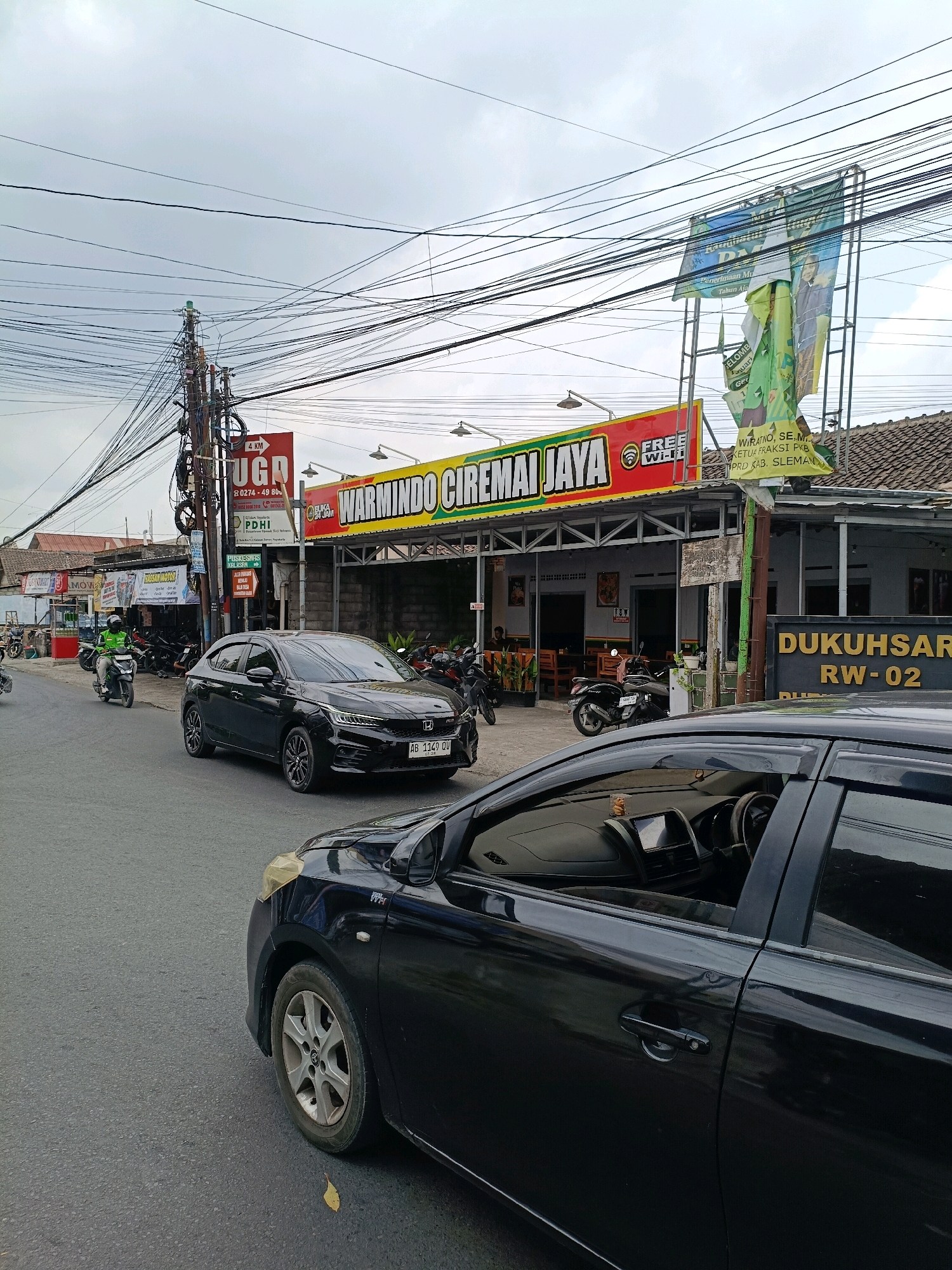 This is a street-level, eye-level, medium-shot photograph taken in Purwomartani, Indonesia. The scene is dominated by a street with two black cars, one in the foreground on the right and another in the mid-ground on the left. In the background, there is a building with a sign that reads "WARMINDO CIREMAI JAYA FREE WI-FI". There are many overhead power lines creating a dense network against a cloudy sky. A motorcycle with a rider in a green jacket is visible on the left side of the street. Several other motorcycles are parked in front of the building. A billboard with an image of a person is visible above the building. The environment appears to be a commercial area with signs indicating services and businesses.