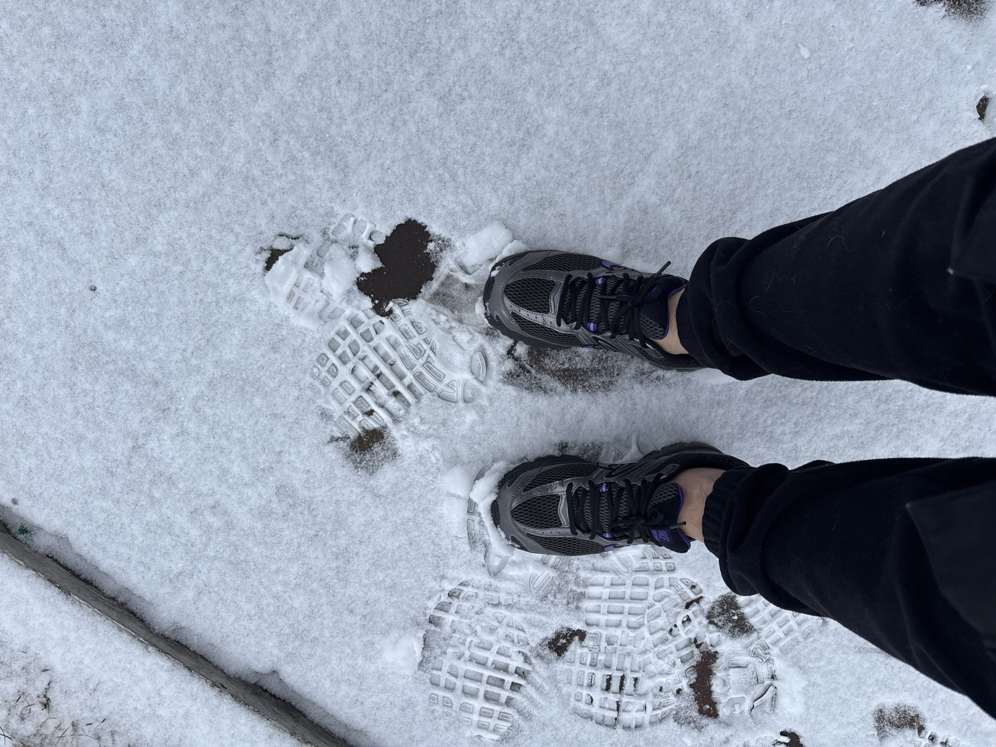 The image shows a person standing in snow. The person is wearing black pants and grey and purple running shoes. There are footprints in the snow, both from the person's shoes and previous prints. There is an area of exposed ground where the snow has been removed. The background is covered in snow. There is a concrete curb at the bottom left. The location cannot be identified as Krasnoyarsk, Russia.