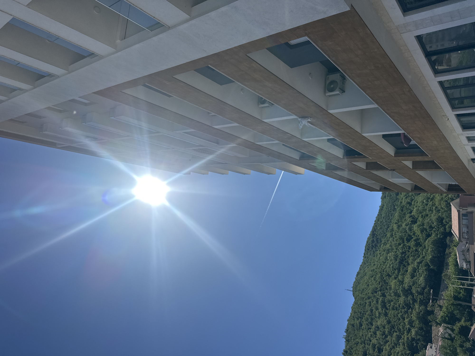 The image depicts a view from below a multi-story building, looking upwards towards a bright sun and a blue sky. The building features a repetitive pattern of projecting rectangular elements, creating a grid-like appearance. A mountain densely covered with trees is visible to the right, and some residential buildings are situated at the base of the mountain. A contrail is visible in the upper portion of the sky. Based on the architectural style and environmental context, this location could possibly be in or near Budva, Montenegro.
