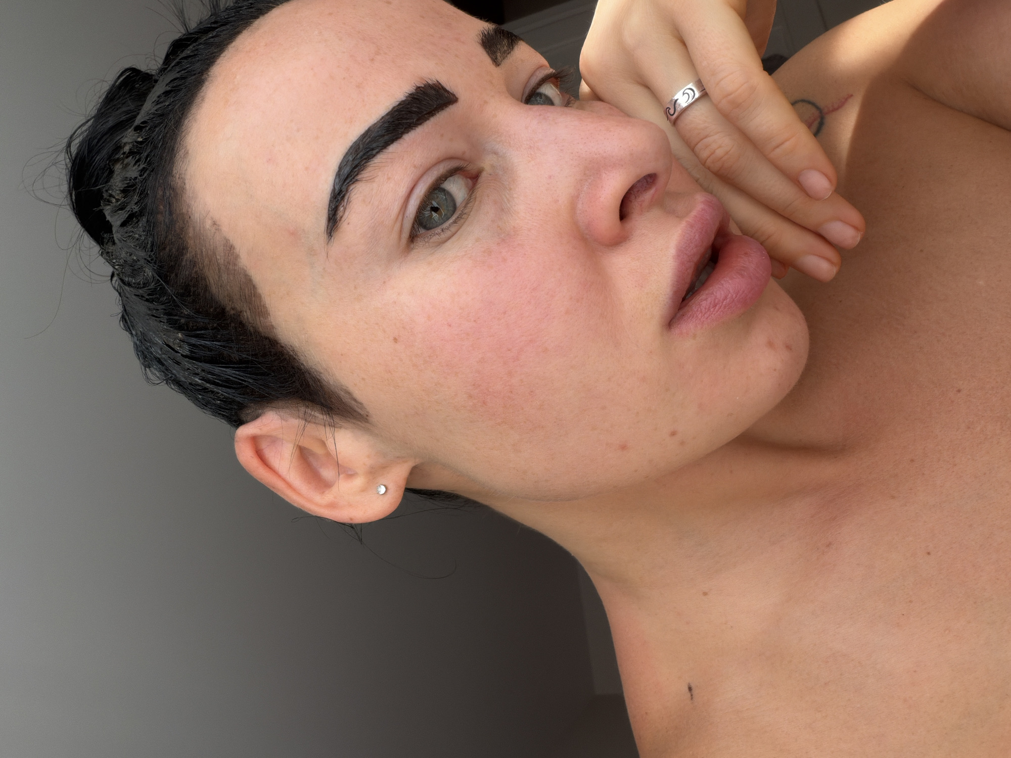 A person is captured in a close-up portrait, seen from the upper chest upwards, with their head tilted and gaze directed towards the camera. Their right hand is raised to their face, with fingers resting gently against their cheek and jawline.
The subject has dark, seemingly wet or styled hair pulled back, exposing their hairline. Their eyebrows are notably dark, thick, and appear to have a freshly applied dark tint or product, with some smudging visible around the brow area, particularly on the left. Their eyes are light-colored, possibly blue or green. Their lips are full and have a natural or light pink hue, slightly parted. The skin tone is fair, with visible freckles or minor blemishes on the face and neck.
On the index finger of their right hand, a silver-toned ring with a distinctive wave or crescent moon pattern is worn. A small, round, silver stud earring is visible in their left earlobe. A faint, circular tattoo or mark is partially visible on the upper right shoulder/collarbone area. A small dark mark is also present on the left side of their neck.
The background is a plain, light grey wall, indicating an indoor setting. The lighting is bright and natural, casting subtle shadows and highlighting features. The overall impression is that of a person engaged in a beauty routine, specifically involving eyebrow tinting, captured in a moment of repose. There is no visible text in the image.