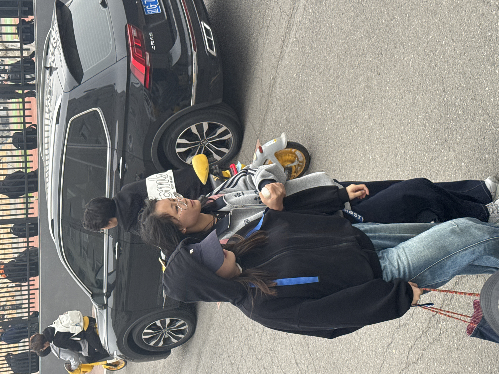 The scene takes place in Zhongtun, China. A black SUV is parked on a grey asphalt surface. A young girl with glasses lies next to the SUV. She has dark hair, wears a grey jacket with black stripes, and dark pants. Next to her, another individual wearing a black hoodie, jeans, and a blue cap is visible. The individual in the black hoodie holds a string, and there is a circular object next to them. Behind the car, a fence can be seen, with bags hanging from it. At a distance, another individual sits on a yellow scooter.