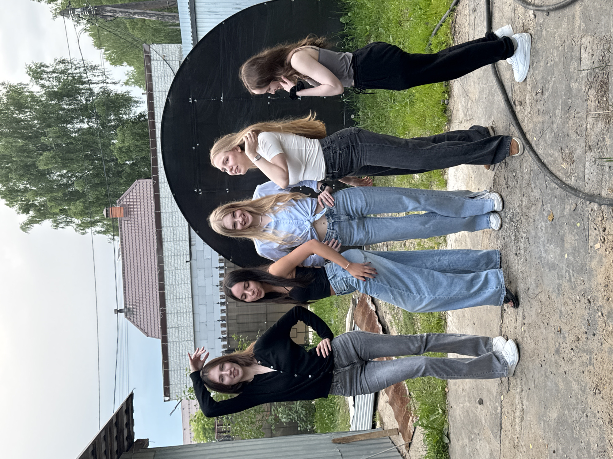 This media file captures a candid outdoor scene featuring five young women posed in a line. The subjects are arranged vertically, with each woman occupying a distinct horizontal space within the frame. They are all dressed in casual, contemporary clothing, including jeans and tops, and appear to be in their late teens or early twenties.
The setting is an outdoor area with a textured concrete ground and patches of grass. In the background, there are glimpses of buildings with pitched roofs and a large, black, circular object, possibly a satellite dish, which serves as a prominent backdrop. Overhead, power lines and lush green trees suggest a semi-urban or rural environment. The sky appears overcast, indicating a cloudy day.
The women are interacting with each other and posing for the camera, with some smiling and others looking more contemplative. Their body language suggests a relaxed and friendly atmosphere, possibly a gathering or a casual photoshoot. One woman at the bottom of the frame is striking a playful pose with her hand raised.
While the specific time of day is not explicitly discernible, the lighting suggests it is daytime. There are no visible text elements within the image. The overall mood is informal and youthful. The location context of Perm, Russia, helps to place the scene geographically but no specific landmarks are identifiable to confirm this.