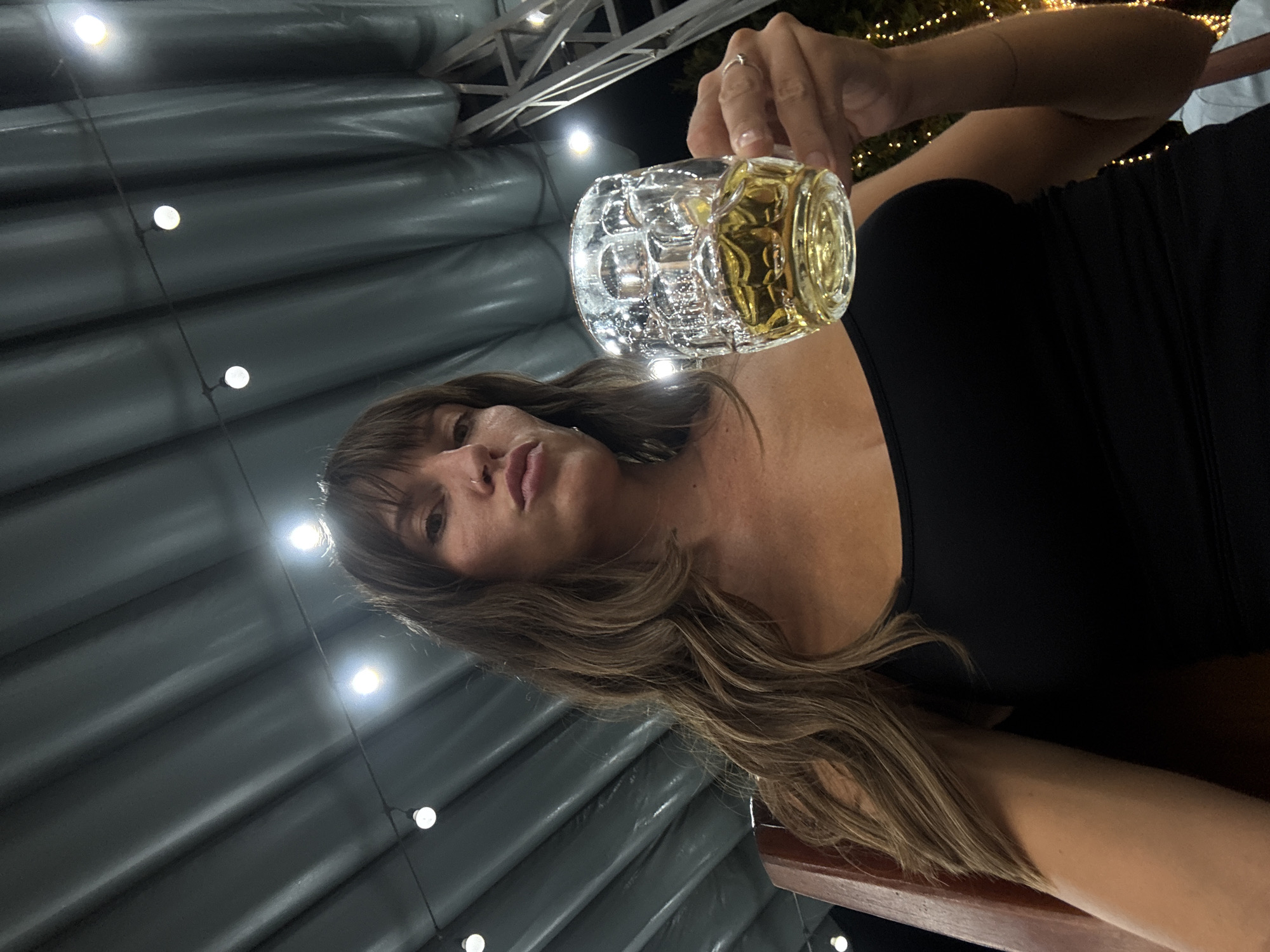 A woman with shoulder-length wavy brown hair and bangs is holding a clear glass mug filled with a yellow liquid, presumably beer. She has fair skin and is wearing a black dress. She appears to be seated at a table. The background features a corrugated wall with evenly spaced light bulbs. The environment suggests an outdoor bar or restaurant. There is no specific evidence in the image to determine the location is Đà Nẵng, Vietnam.