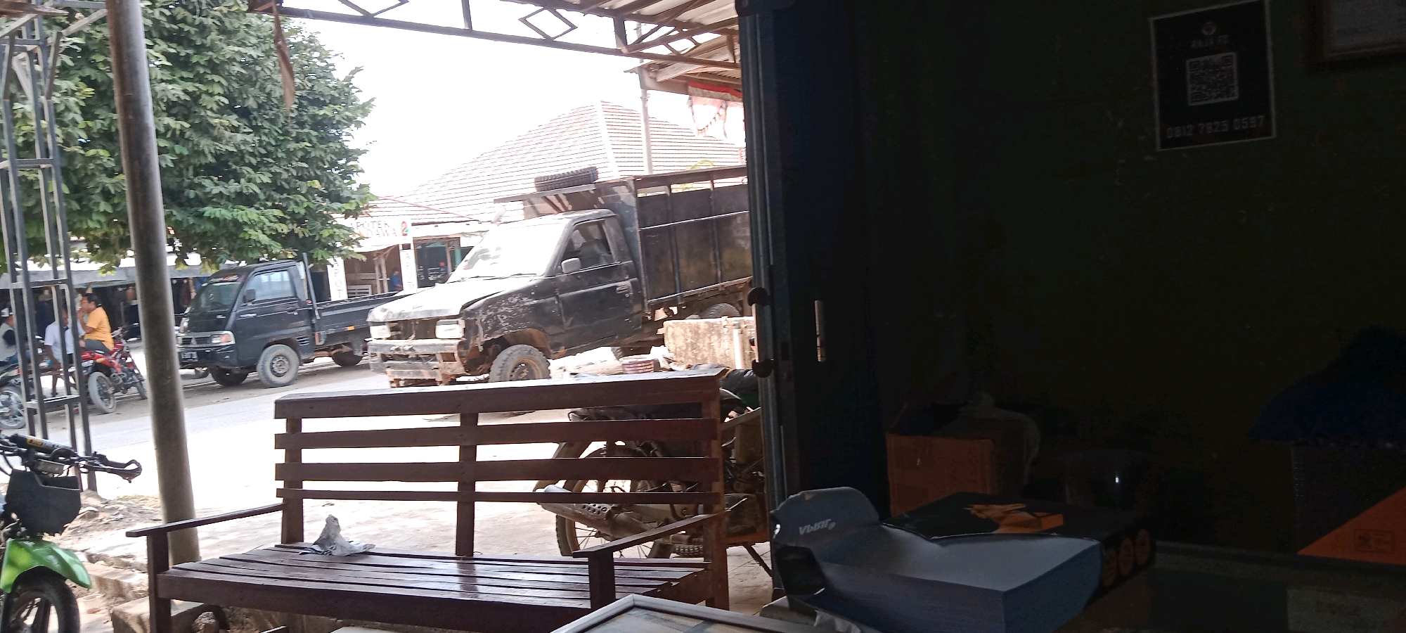 The image depicts a street scene in Binakarya Putra, Indonesia, likely in front of a workshop or garage. In the foreground, a wooden bench is visible, with a green motorcycle parked to its left. Behind the bench, a black truck with a dilapidated front end and a spare tire on its roof is parked on the street. Another black pickup truck is parked to the left of this truck. A building with a white facade and a tiled roof is visible in the background, with trees and other structures lining the street. The scene appears to be outdoors, under what seems to be a covered area. The overall impression is one of a busy street or commercial area, with vehicles and buildings indicative of a working-class environment.