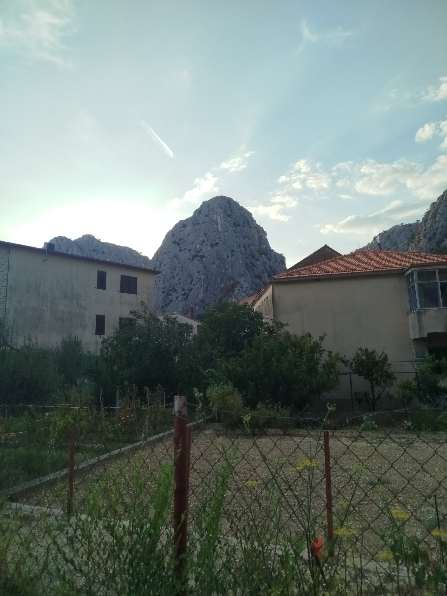 The image captures a residential area in Omiš, Croatia, with a backdrop of imposing rock formations. In the foreground, a chain-link fence encloses a garden area with some vegetation. Beyond the fence, two buildings are visible. The building on the left is a multi-story structure with a light-colored facade and several dark-framed windows. The building on the right features a terracotta-tiled roof and a similar light-colored facade, with an enclosed balcony or sunroom. Lush green trees and bushes surround the buildings and gardens, creating a contrast with the rugged mountains that dominate the background. The sky above is a pale blue, with scattered clouds and a faint contrail from an aircraft. The lighting suggests it is either early morning or late afternoon.