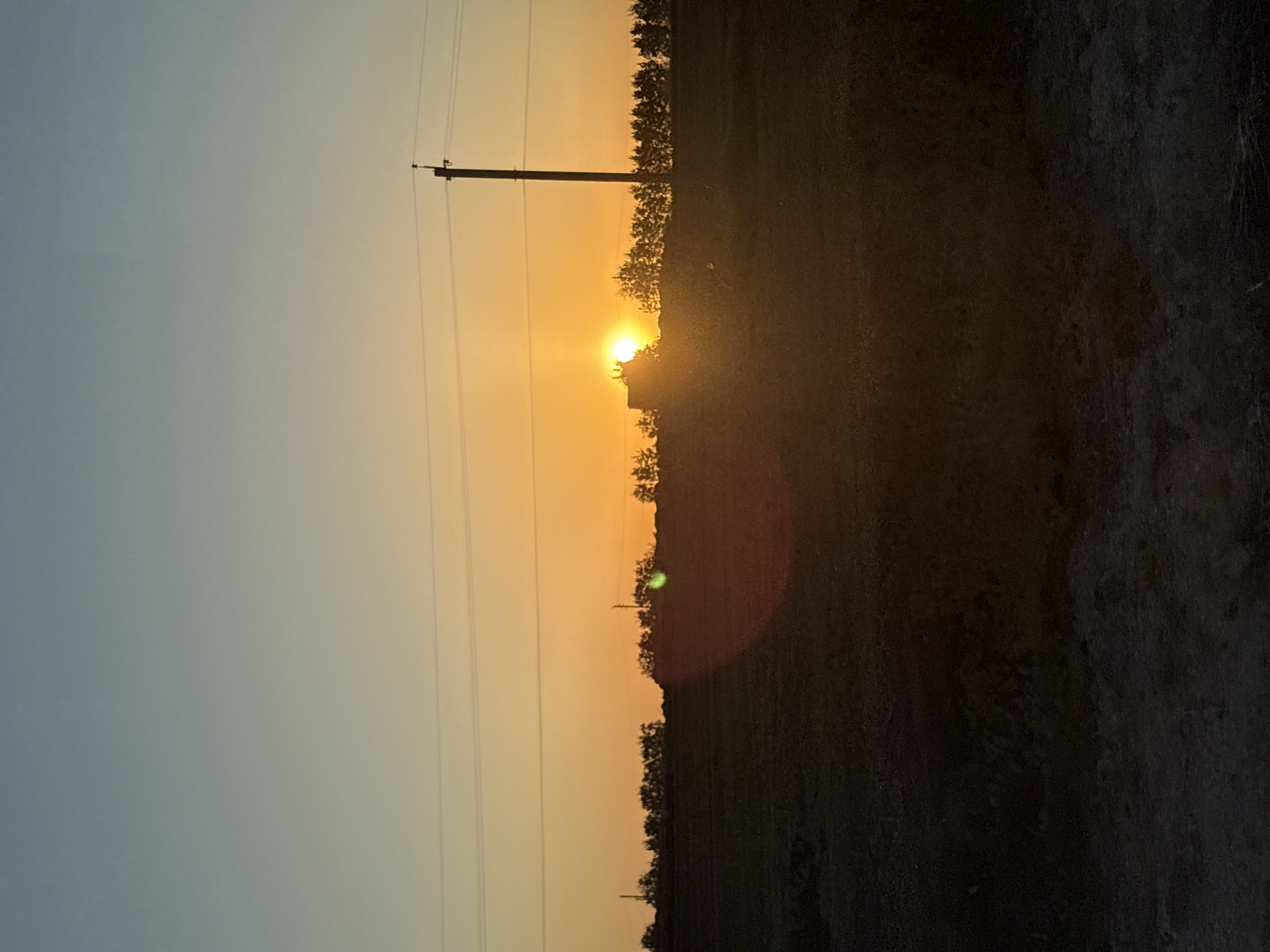 The image depicts a rural landscape at sunset in what could be Bol'shiye Klyuchishchi, Russia. A utility pole stands to the left of the sun, with wires stretching across the scene. Silhouetted trees and vegetation form the horizon line, obscuring the setting sun. A lens flare is visible. The foreground is dark and indistinct. The sky transitions from blue to orange, indicating sunset. The image has been taken with a camera that has rendered the image vertically.