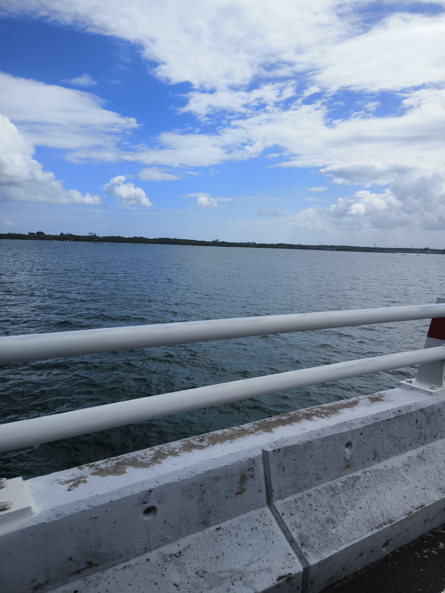 The image depicts a tranquil scene of a body of water, likely a bay or a wide river, under a partly cloudy sky. The water is a deep blue-green with gentle ripples, suggesting a calm day. In the foreground, a white railing runs horizontally across the image, partially obscuring the view of the water. This railing appears to be part of a bridge or pier, with a concrete barrier visible below it. The concrete barrier is weathered and shows signs of age, with patches of discoloration and some staining.
The far bank of the water is lined with lush green trees, forming a natural horizon. In the distance, faint structures and what appear to be power lines can be seen, hinting at a developed area beyond the natural landscape. The sky is a vibrant blue, punctuated by fluffy white clouds that cast soft shadows on the water. The overall impression is one of peacefulness and natural beauty, with a subtle hint of human infrastructure. This scene is located on the southern coast of Bali, in the district of Kuta Selatan, Indonesia.