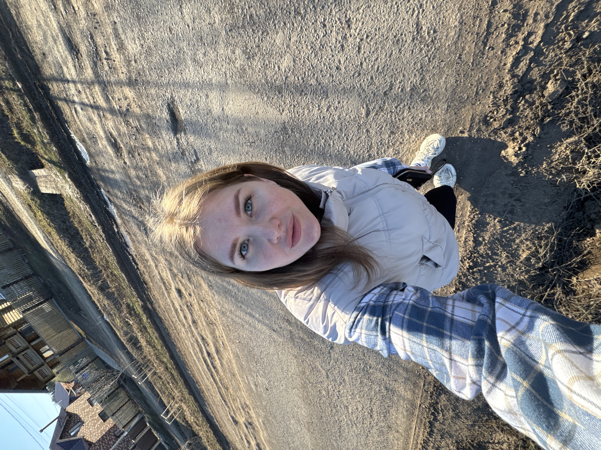 A young woman with light brown hair, blue eyes, and light skin is taking a selfie outdoors. She is wearing a light beige puffer jacket and a blue and white plaid flannel shirt. Her white sneakers are visible. The background shows a dirt road or path with grassy areas on either side. In the distance, there are buildings, potentially houses, under a clear sky. The scene appears to be Горный, Russia, based on visual indicators like the style of buildings. The woman is facing the camera directly, holding the phone out at arm's length to capture the selfie.