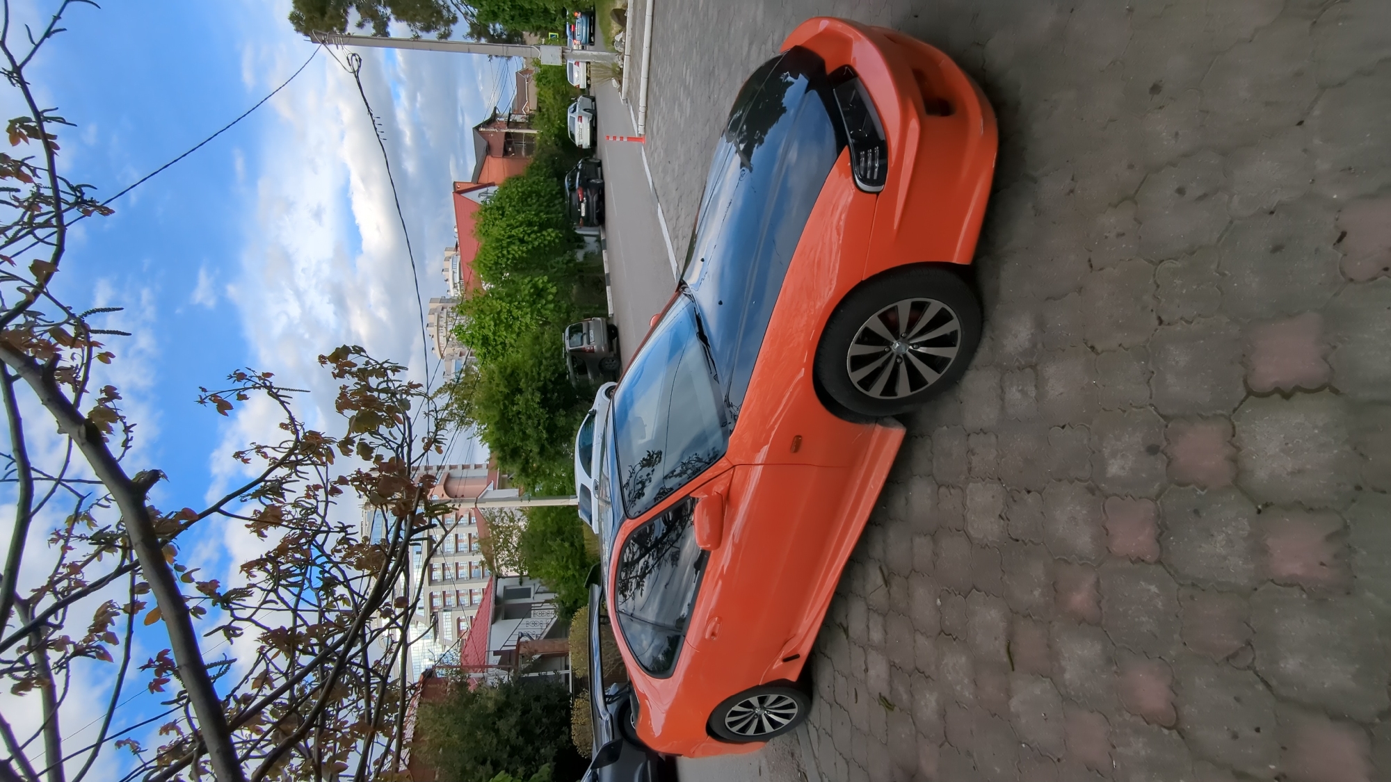 An eye-level, rotated, wide-angle shot shows an orange 1990s Honda CRX sports car parked on a patterned gray brick driveway in Gelendzhik, Russia. The car has a black top. The background shows a city landscape including trees, other parked cars, and various buildings with red roofs. The shot includes a foreground of tree branches on the left and a blue sky in the upper left corner.
