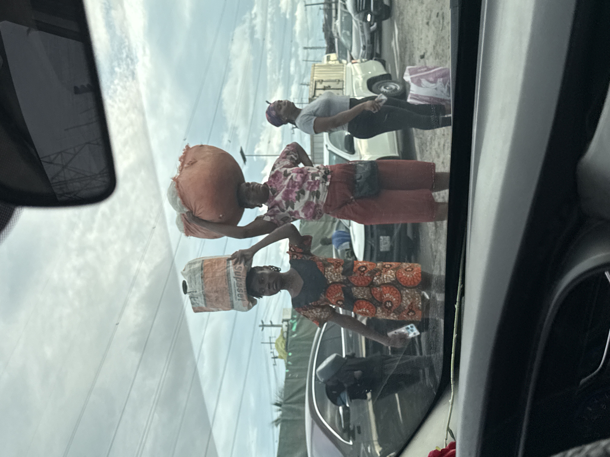 The image, taken from inside a car, shows three women outside the vehicle, with cars and infrastructure visible in the background. The woman closest to the camera is carrying a bag of "Supaset" on her head and holding a smartphone in her right hand. She is wearing an orange and brown patterned dress. Behind her, another woman is carrying a large, round orange bag on her head and wearing a floral shirt and red pants. A third woman, wearing a gray shirt and black pants, is standing behind them. The scene appears to be in Igboefon, Nigeria, based on the "Supaset" bag which suggests a local cement brand. Other cars are visible in the background, indicating a street or roadway. Power lines are also visible overhead. The view is partially obscured by the car's windshield and interior components, including the rearview mirror.