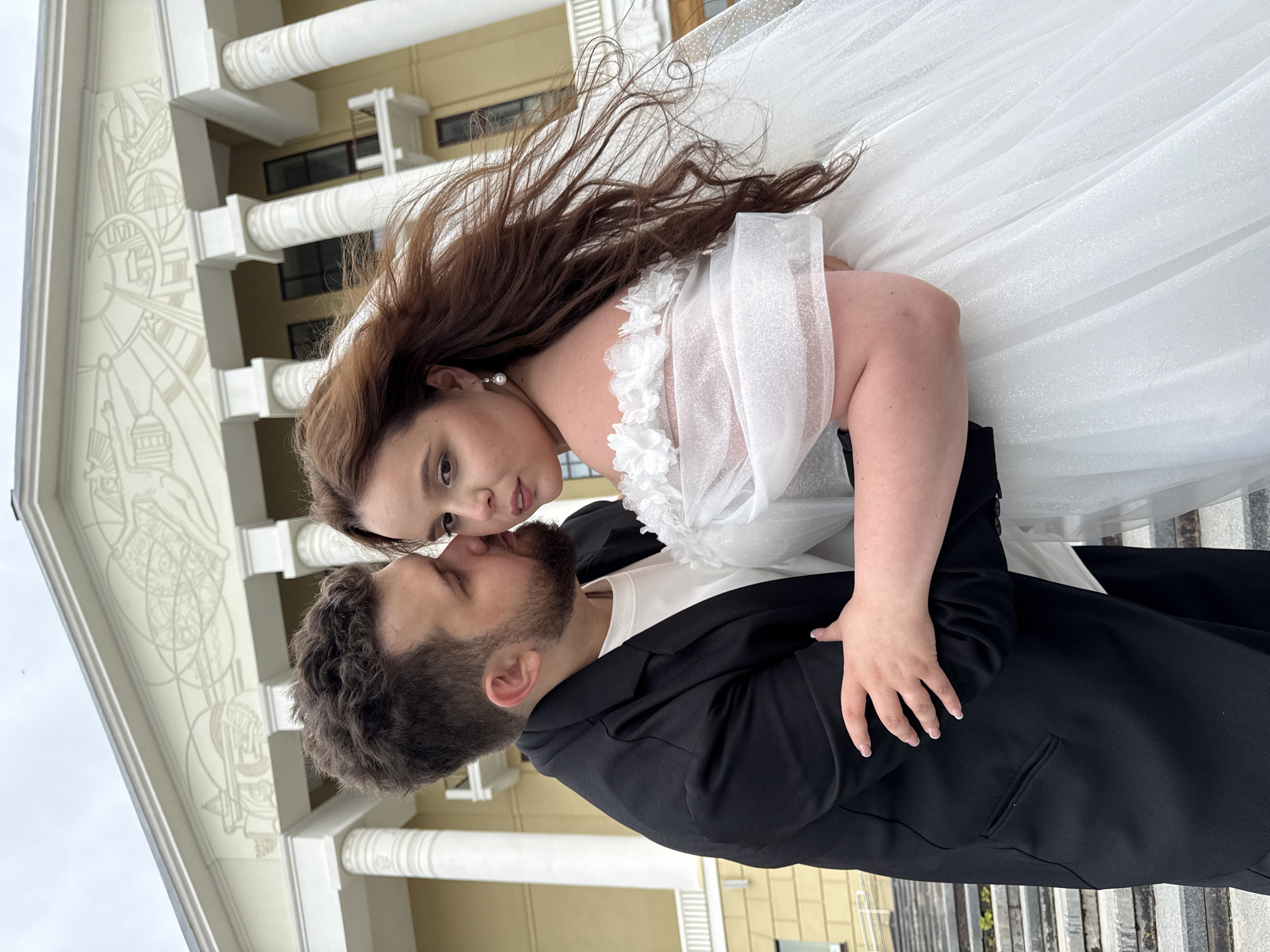 The image shows a bride and groom in front of a building with large white columns. The groom, on the left, is wearing a black suit jacket and a white shirt. He has his arm around the bride and is whispering in her ear. The bride, on the right, is wearing a white wedding dress with off-the-shoulder sleeves and pearl earrings. Her hair is long and brown and appears to be blowing in the wind. The background building appears to be a significant public building, possibly a cultural center or museum, with neoclassical architecture. The sky is overcast. The location is the Votkinsk State Museum-Reserve, a cultural heritage site in Votkinsk, Russia, known for its association with Pyotr Ilyich Tchaikovsky. The couple is positioned on stone steps leading up to the building.