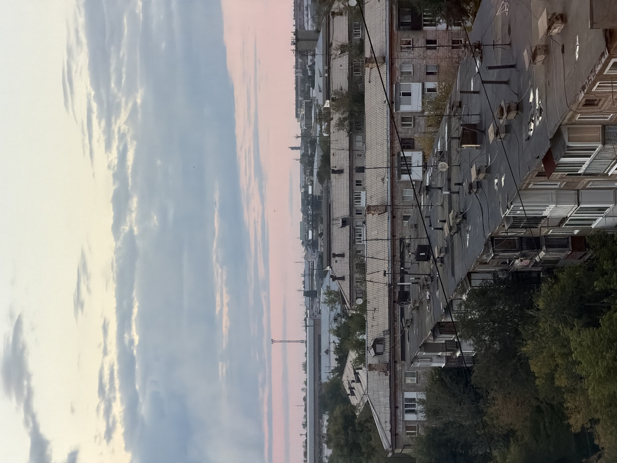 The image depicts a cityscape, likely in Pavlodar, Kazakhstan, characterized by overcast skies with hues of pink and pale yellow. Dominating the mid-ground are rows of Soviet-era apartment buildings, their flat, dark roofs adorned with various vents, chimneys, and antennas. The architecture is consistent with the typical panel buildings found in post-Soviet urban areas, featuring repetitive window patterns and enclosed balconies. Green trees, some with autumn foliage, are interspersed between and around the buildings, suggesting an urban setting with pockets of natural growth. In the distance, industrial structures and a prominent radio tower are visible, hinting at the city's industrial character. The overall impression is one of an everyday urban scene, potentially captured during twilight or a cloudy day.