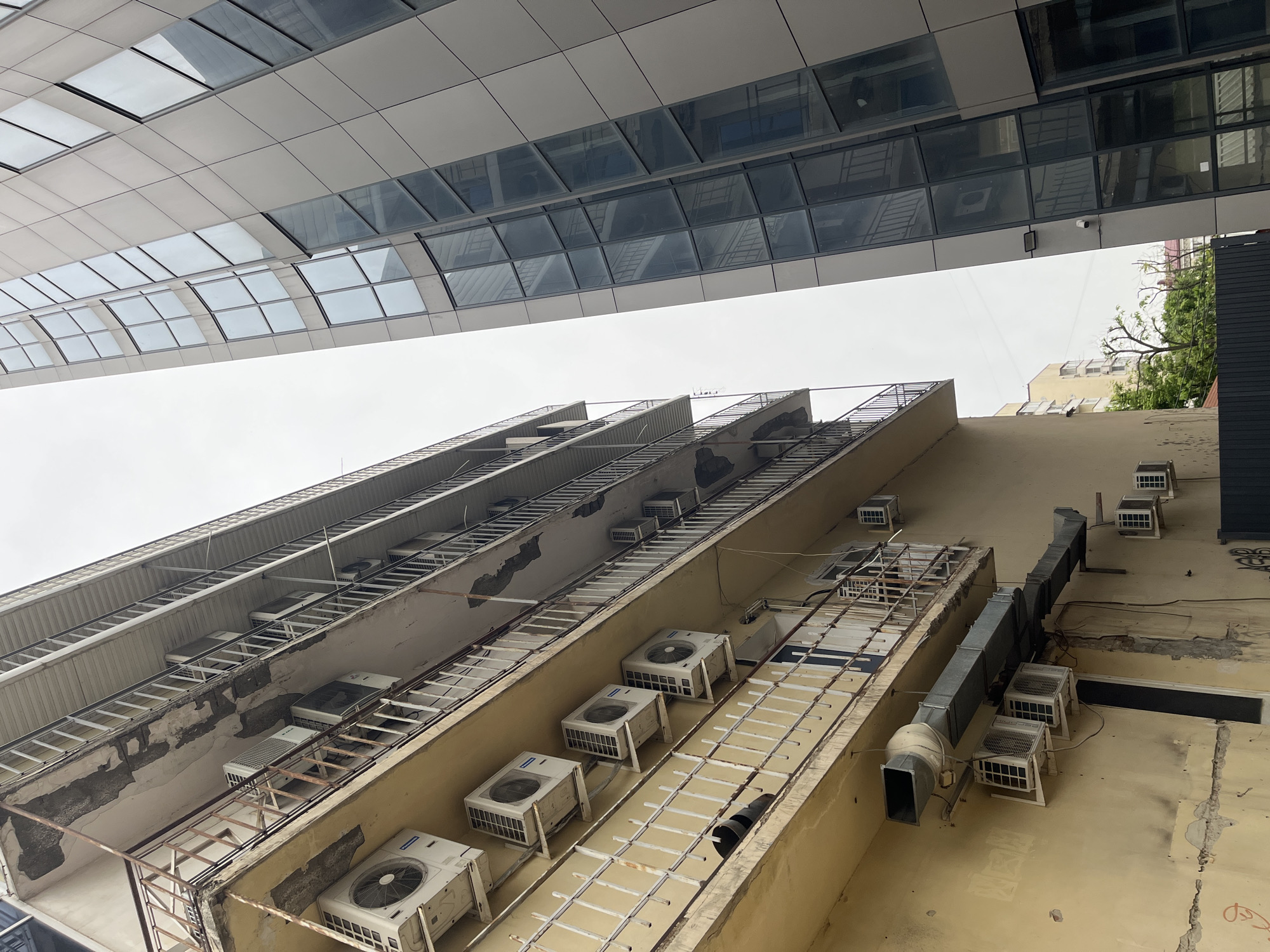 The image shows an upward view of the exterior of buildings in an urban setting. A modern building with a glass facade and metal paneling is visible at the top, contrasting with the older, weathered building below. The older building has multiple levels, with a series of air conditioning units installed along its facade. Metal ladders and framework are attached to the building, likely for maintenance access. The color palette is dominated by whites, grays, and yellows, giving a slightly industrial feel. The sky is overcast and visible between the buildings. The image does not provide conclusive visual information to confirm the location in Krasnodar, Russia. The presence of air conditioning units and the architectural style suggest a temperate or warm climate. The scene lacks people, subjects, actions, or interactions. The focus is on the architectural details and the built environment.