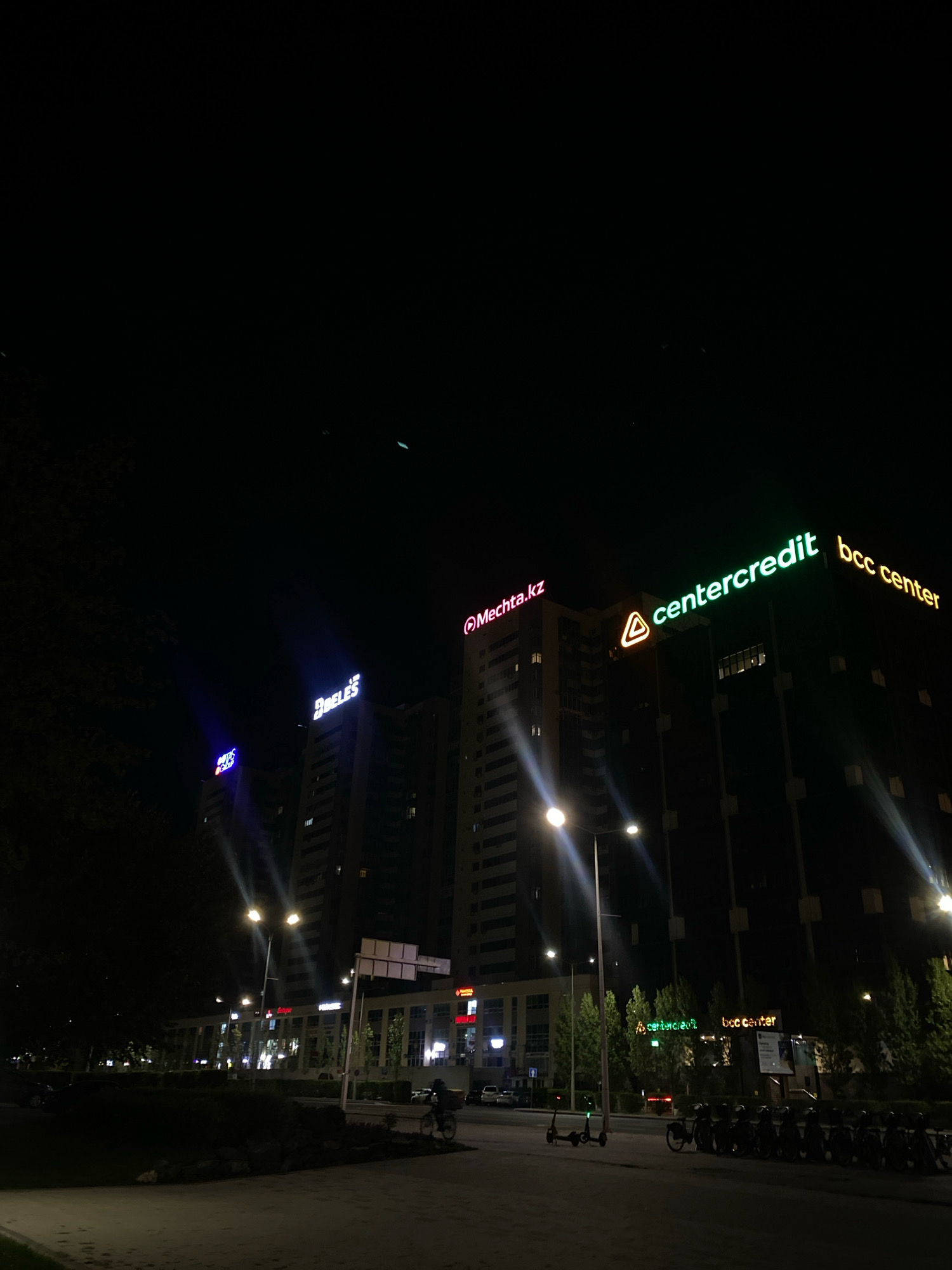 In Astana, Kazakhstan, a nighttime scene unfolds along a city street. The foreground features a paved walkway bordering a dark, grassy area with scattered rocks. Beyond this, a street is lined with lampposts and vehicles, including parked cars and a row of bicycles. In the middle ground, a low-rise building with illuminated windows and signage stretches across the scene.
Dominating the background are several tall buildings. The building on the right displays prominent illuminated signage that reads "centercredit bcc center" in green and yellow. To its left, another building features a pink neon sign with the text "Mechtа.kz" and a triangular logo. Further left, a building shows blue illuminated signage with the text "BOTOS" and another smaller word, possibly "CREDIT". The overall atmosphere is dark, with artificial lights from the buildings and streetlights illuminating the urban landscape.