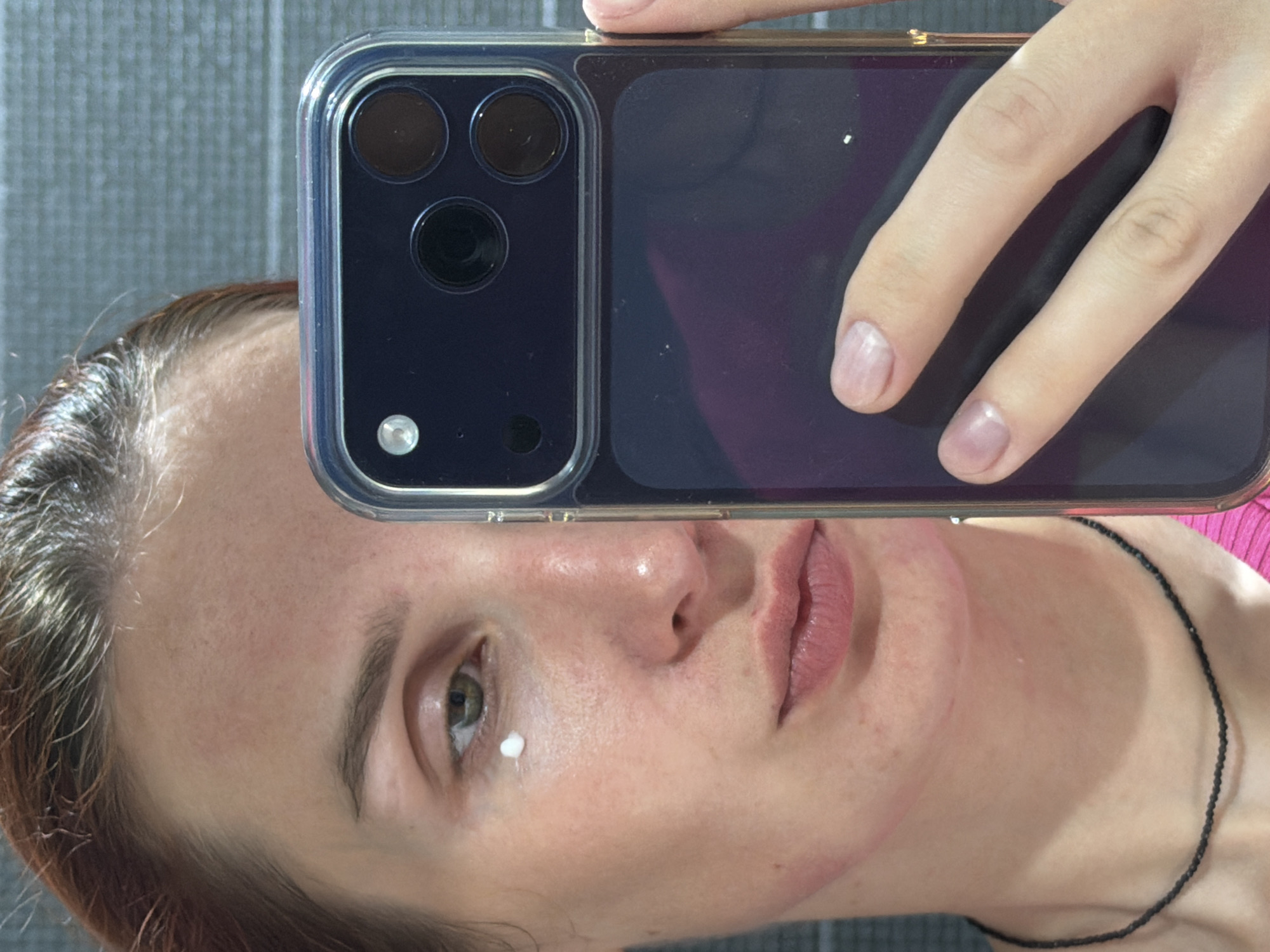 A woman is taking a selfie with a smartphone. The phone partially obscures her face. She has fair skin and light brown hair. A small amount of white cream or lotion is present near her left eye. She is wearing a black beaded necklace. The background appears to be a gray textured surface. There is no identifiable indication of the city of Chaikovsky, Russia.