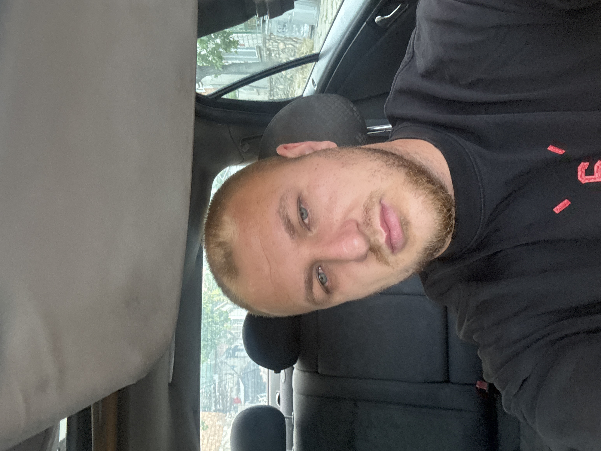 An image is presented, rotated 90 degrees counter-clockwise, showing a man inside what appears to be a car.
**Subjects:** The primary subject is a man with short, light-colored hair and a light-brown beard/stubble. He has light-colored eyes (blue or greenish) and pink lips. He is wearing a black, long-sleeved shirt or sweatshirt.
**Scene/Setting:** The man is situated in the interior of a vehicle, likely a car, with a dark headrest visible behind him and the car's headliner in the upper portion of the frame. Through the car window, an outdoor environment with trees and what appear to be buildings or structures can be seen, though somewhat distorted by the glass.
**Activity or event taking place:** The man is looking directly at the camera with a neutral to slightly relaxed expression, suggesting he is posing for a photo or selfie.
**Notable details:**
* **Time of day/Weather:** The lighting from outside the window indicates it is daytime, possibly overcast or bright.
* **Location cues:** The visible car interior components and the background suggest an urban or suburban setting, though no specific geographical landmarks are identifiable.
* **Emotions:** The man's expression appears calm and composed.
* **Orientation:** The image is captured and displayed rotated on its side.
**Visible text:** On the man's black shirt, there are red markings that resemble parts of stylized letters or a logo, specifically two vertical red dashes and what looks like the top-left curve of a letter "J" or similar character.