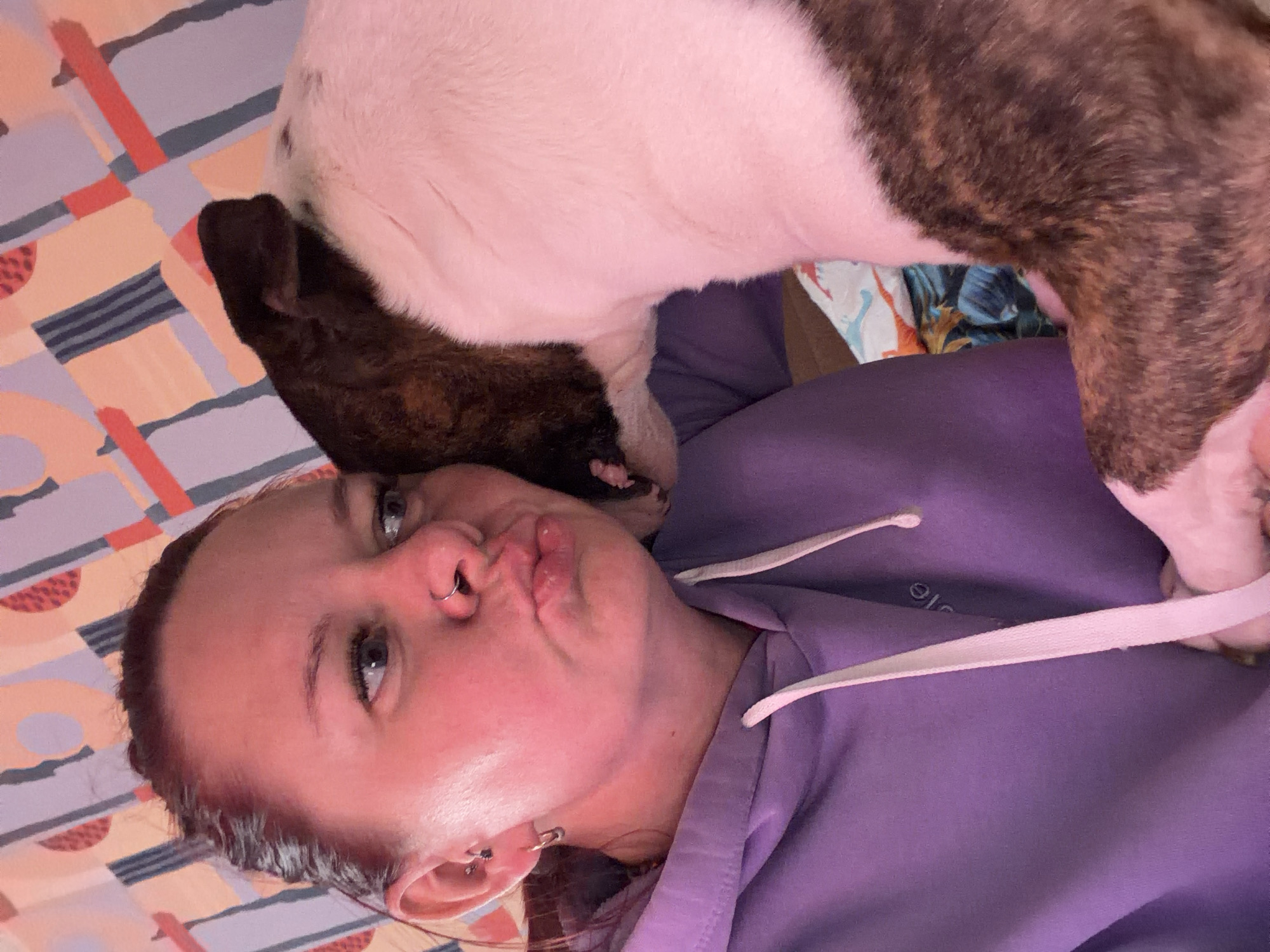 A person with light skin and red hair, wearing a purple hooded sweatshirt, is lying on a patterned surface. Their lips are puckered. A brindle and white dog is standing on top of them, with its head and front paws resting on the person's face and chest. The background features a colorful patterned surface. It is impossible to determine from the image whether the location is Chaikovsky, Russia.
