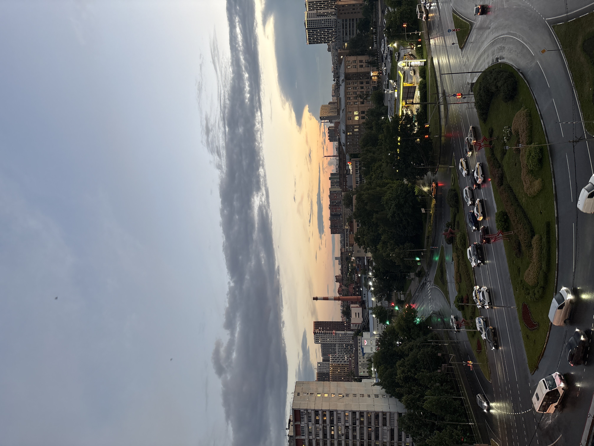 This is a high-angle, slightly rotated, wide shot of a city intersection during dusk. The sky is a mix of light grey clouds and a warm orange and yellow sunset. Tall buildings, some with illuminated windows, line the sides of the road. Trees are also visible, creating a green canopy.
The main focus is a multi-lane road with several cars driving on it. The road appears wet, suggesting it has recently rained or is still raining lightly. Traffic lights are visible, and some are red, indicating traffic is stopped. A circular traffic island with manicured bushes and trees is at the center of the intersection. There is a prominent red star-shaped structure on the traffic island.
In the distance, a tall brick chimney with a red cap stands out against the skyline. The overall mood is calm and serene, with the soft lighting of dusk and the quiet hum of the city. The location is identified as Chelyuskinsky, Russia.