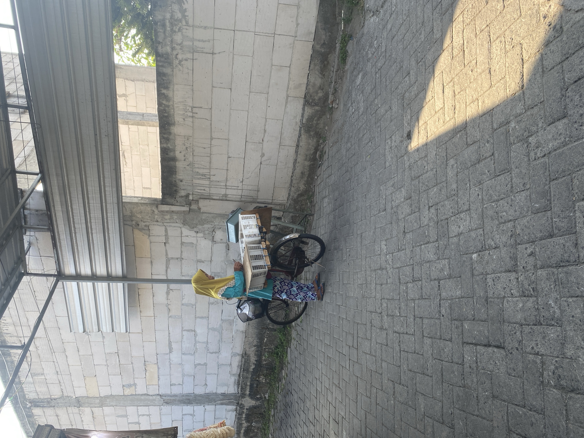 A woman in a yellow hijab and a blue and patterned outfit stands next to a bicycle with a basket on the front. The bicycle is loaded with several white crates. She is looking down at her phone. The bicycle is parked on a grey brick pathway. Behind the woman is a white brick wall. Above the wall is a corrugated metal roof supported by metal beams. The scene appears to be outdoors, possibly in a market or alleyway. The lighting suggests it is daytime with sunlight casting a shadow on the pathway. The location is specified as Kediri City, Indonesia.
