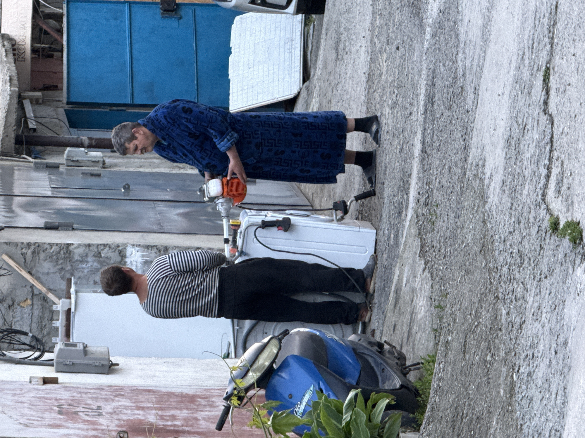 The image shows two people outside in what appears to be an urban environment. One man is wearing a blue robe with a pattern on it and black socks. He is holding an orange and white machine. The second man is wearing a striped shirt and black pants. Next to him is a white object with electrical cords. A blue and black scooter is parked in front of the white object. The ground is concrete. The background consists of buildings with metal garage doors.