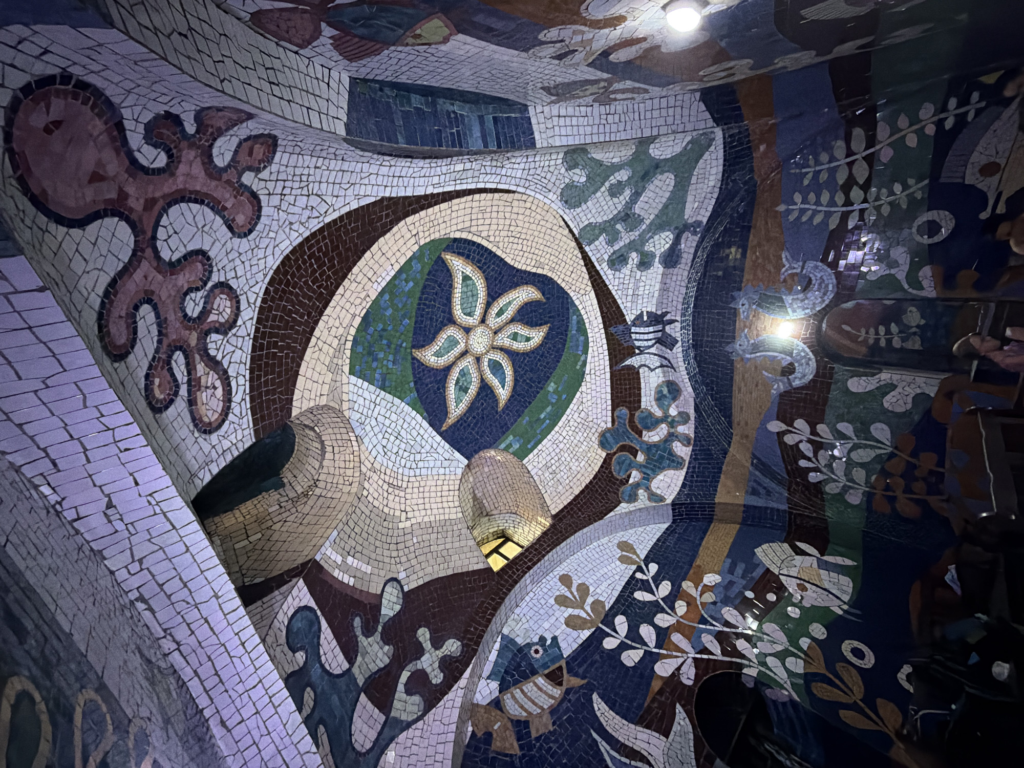 This image depicts a detailed mosaic art installation, likely on a curved surface such as a wall or ceiling. The mosaic features various motifs including floral patterns, fish, birds, and abstract shapes, all rendered in a variety of colors. The work appears to have been executed with small, square mosaic tiles of varying shades and hues. The installation is lit by a light source, which creates highlights and shadows on the mosaic surface.
This is likely a part of the mosaic artwork located in the Tbilisi Public Service Hall in Tbilisi, Georgia.
