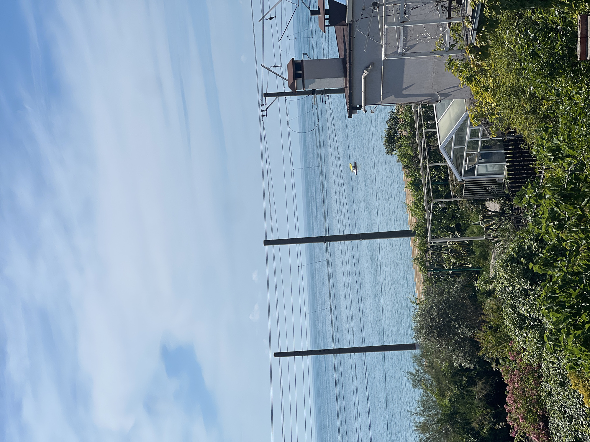 The image shows a view of the sea from a garden area. In the foreground, there is lush green vegetation, including bushes and flowering plants. A greenhouse is visible, along with a portion of a house with a chimney. In the mid-ground, there is a section of beach and the calm blue sea. Several vertical poles with overhead wires run across the scene, suggesting the presence of a railway or power lines. In the distance, on the water, a small yellow boat is visible. The sky is blue with scattered white clouds. The location is described as Unknown, Unknown.