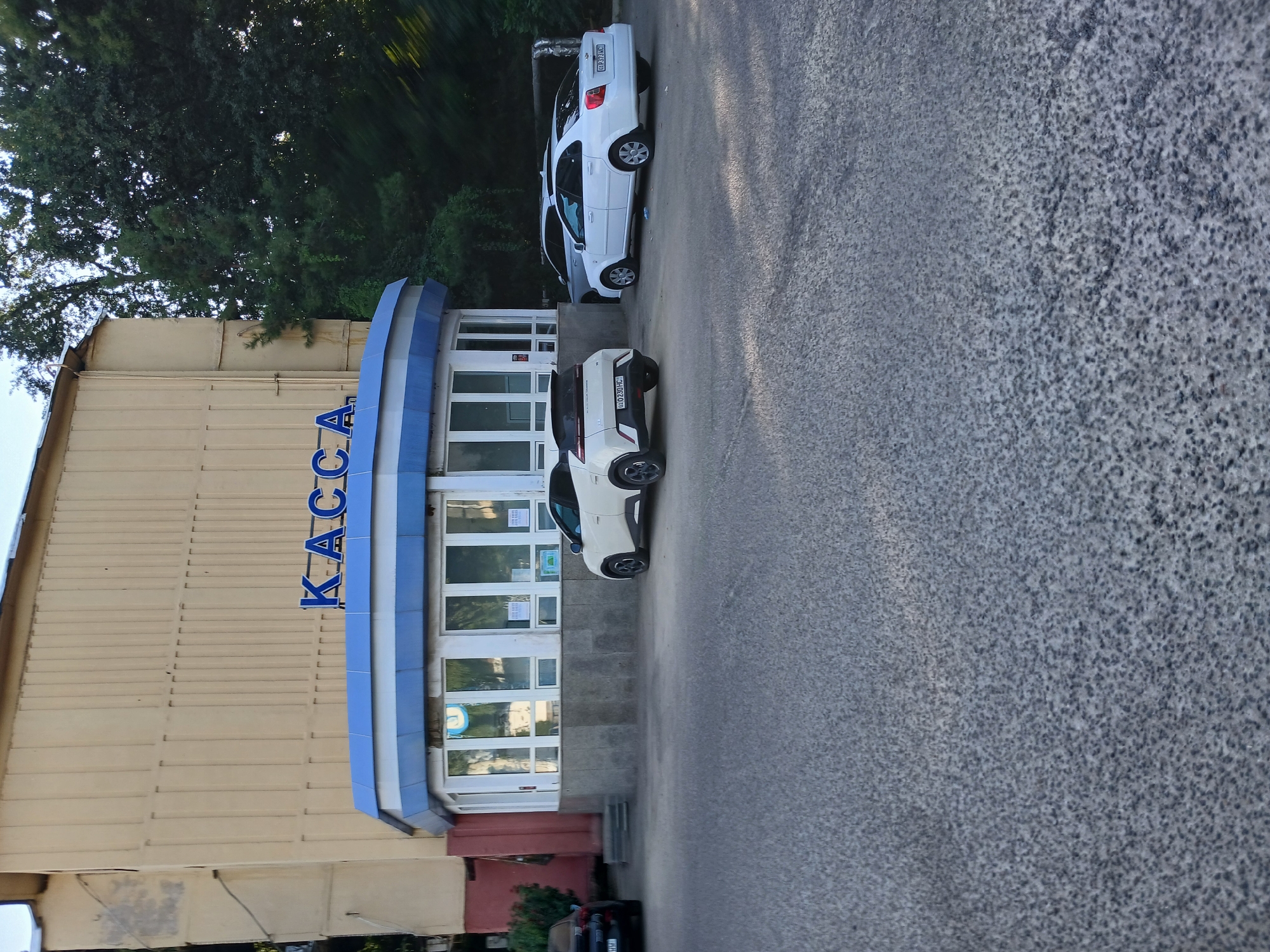 A daylight exterior shot captures a light-yellow, three-story building with vertical ribbed siding in Tashkent, Uzbekistan. Above a rounded, blue-and-white covered entrance with large windows, the Russian word "КАССА" (KASSA), meaning "cash desk" or "ticket office," is displayed in blue capital letters. The entrance area is adorned with multiple windows and a textured gray stone base. In front of the building, parked on an asphalt surface, are three cars: a black sedan on the far left, a white hatchback in the middle, and a white sedan on the right. Lush green trees are visible in the background, casting shadows on the scene.