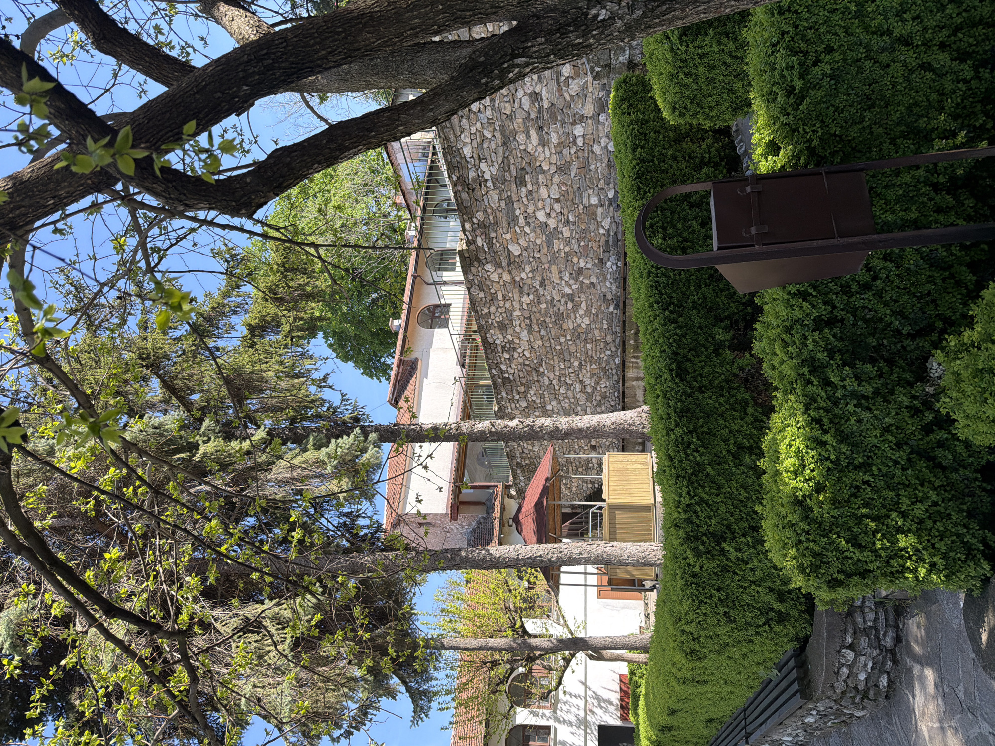 The image shows a building in Brestnik, Bulgaria, viewed through trees. The building has a stone facade and multiple stories, with visible balconies and windows. A tall, manicured hedge runs along the right side of the image, partially obscuring a metal structure resembling a cart. A stone wall is visible beneath the hedge. Branches and leaves from trees frame the view on the left and top of the image, with a clear blue sky visible in the background. Other houses with terracotta roofs are also visible in the background.